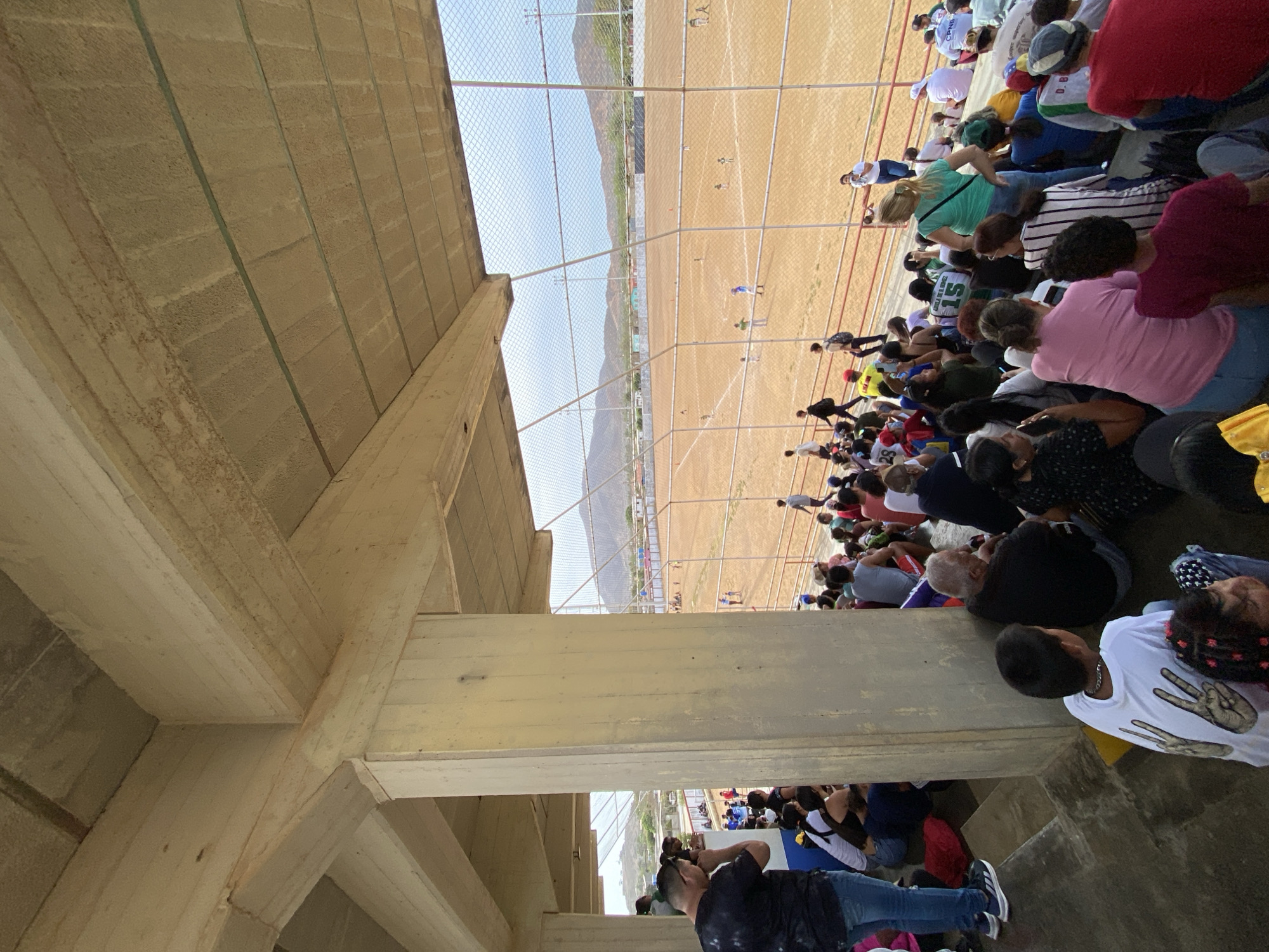 The image depicts a crowd of people gathered at a sports field in Juan Griego, Venezuela. The spectators are seated on concrete bleachers under an overhanging structure. A chain-link fence separates the crowd from the dirt field where athletes are practicing track and field, including running. There are mountains in the background. The environment is sunny.