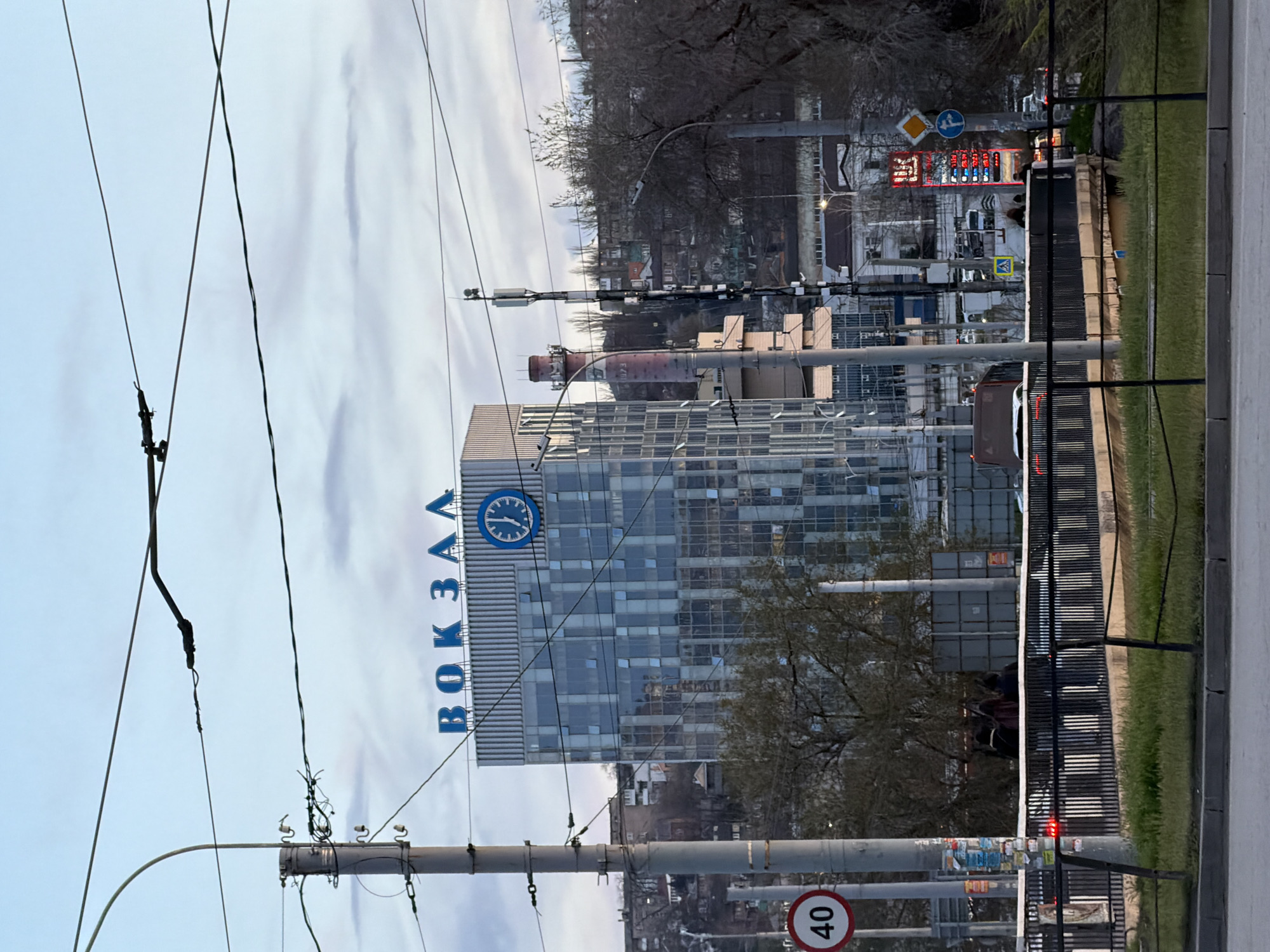 A street scene in Rostov-on-Don, Russia, featuring the Rostov-Glavny railway station. The train station building, with a clock and Cyrillic letters spelling "вокзал" (railway station), is in the background. Overhead power lines and utility poles run along the street, partly obstructing the view of the building. A speed limit sign with "40" is visible near the bottom of the frame. In the background, there are other buildings, including one with a price display, likely for a gas station, and some streetlights. A metal fence and some greenery are present along the edge of the street.