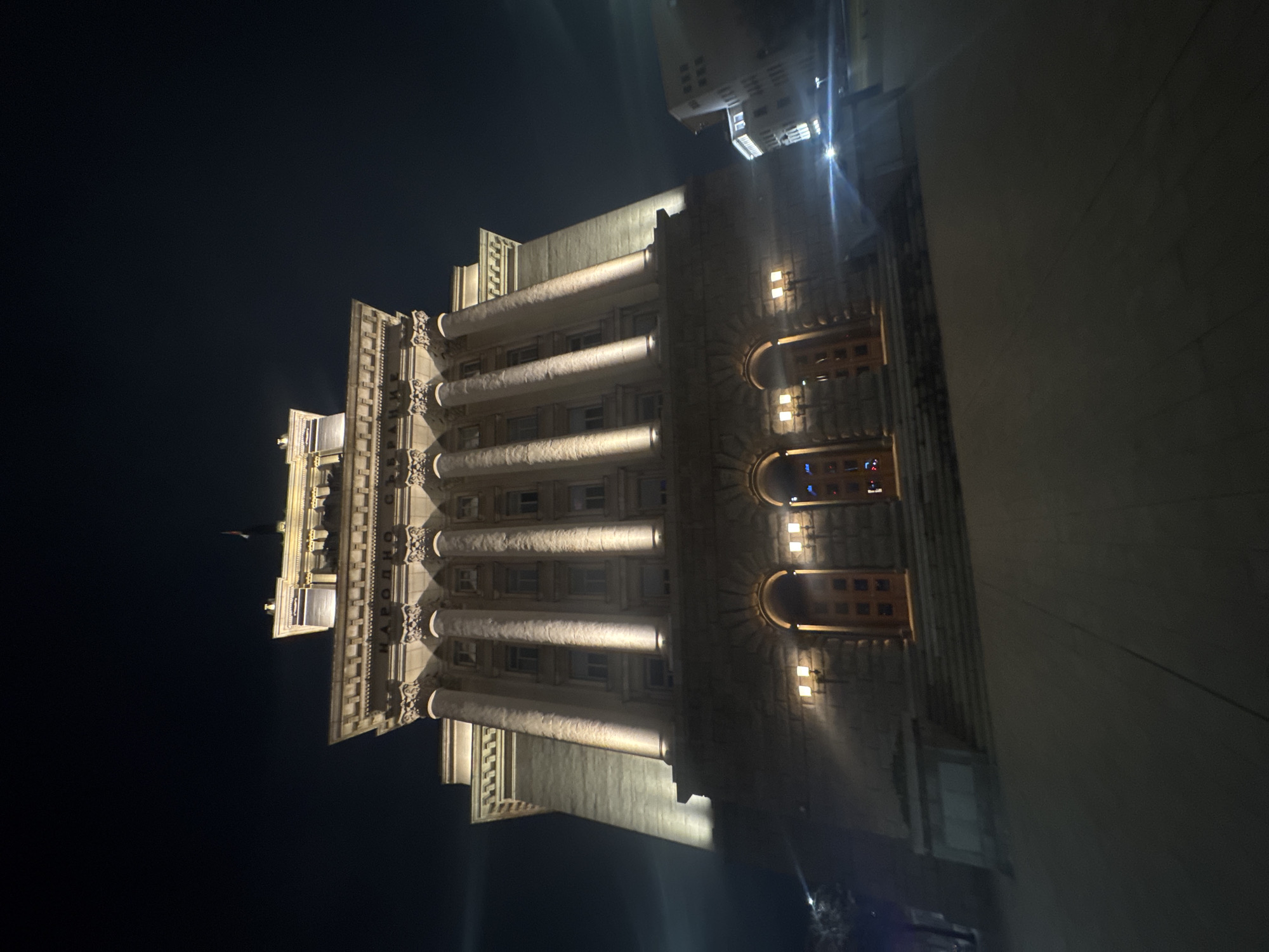 The image shows the National Assembly of Bulgaria building at night in Sofia, Bulgaria. The building is illuminated, highlighting its neoclassical architecture with columns and ornate details. The night sky is dark, and the building stands out against it. Adjacent areas are less illuminated.