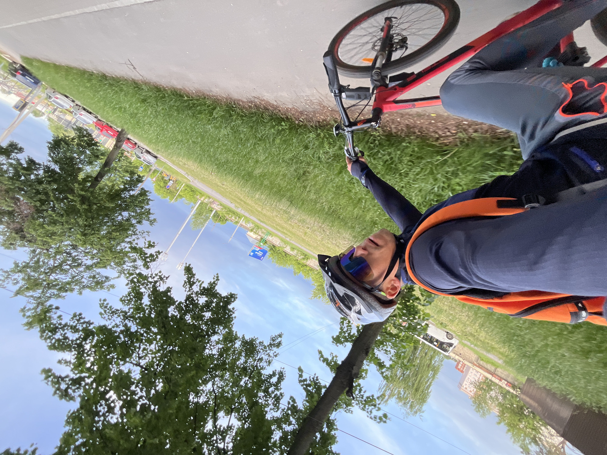 A cyclist is riding a red bicycle on a path in Chaikovsky, Russia. The cyclist is wearing a helmet, sunglasses, a dark blue long-sleeved shirt, gray pants, and an orange safety vest. The bike path runs alongside a grassy area and a road with parked cars and buildings in the background. Trees line the path, and the sky is blue with some clouds. A white bus is visible further down the path. The image appears to have been taken as a selfie by the cyclist.