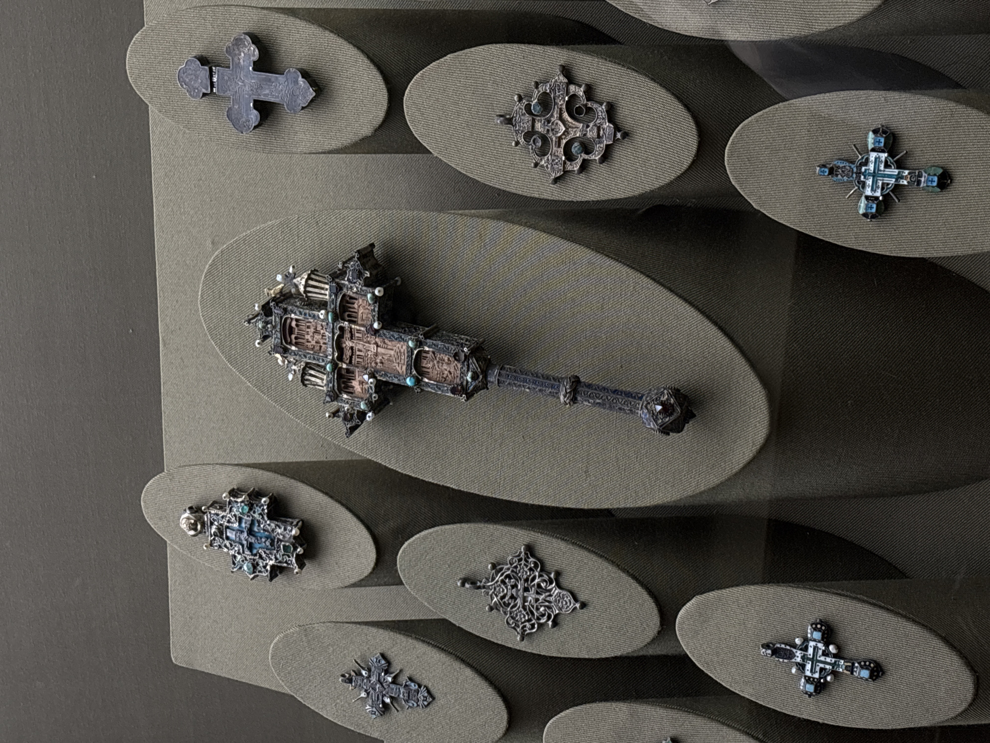 The image depicts a collection of historical artifacts, likely religious objects, displayed on a textured, olive green fabric backdrop. The artifacts, primarily crosses of varying designs, are arranged on individual oval platforms, also covered in the same fabric. One object stands out due to its larger size and unique shape: a mace or scepter-like item with a decorative head and long handle. The artifacts appear to be made of metal, some embellished with stones or enamel. The setting suggests a museum display, possibly in a city like Moscow, Russia, known for its rich history and religious heritage. No people are visible.