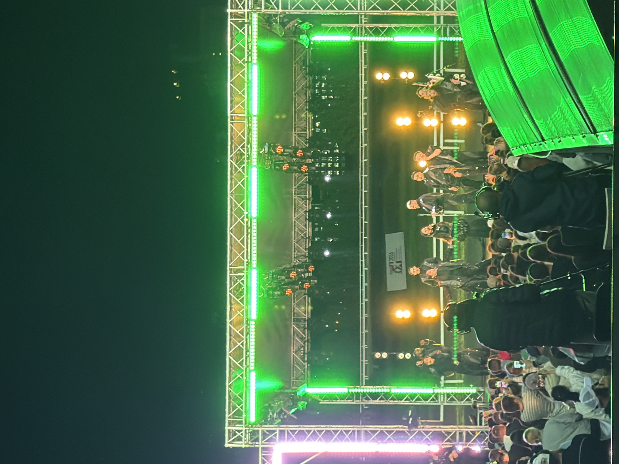 An outdoor nighttime concert is underway, with a stage illuminated by bright green lights. A group of performers, dressed in black, stand on the stage. In front of the stage, a large crowd of spectators is gathered. Some audience members are holding up their phones, presumably to take pictures or videos. A large, curved green structure, possibly a speaker or lighting fixture, is positioned in the foreground on the right side of the image. The background is dark, indicating it's nighttime.