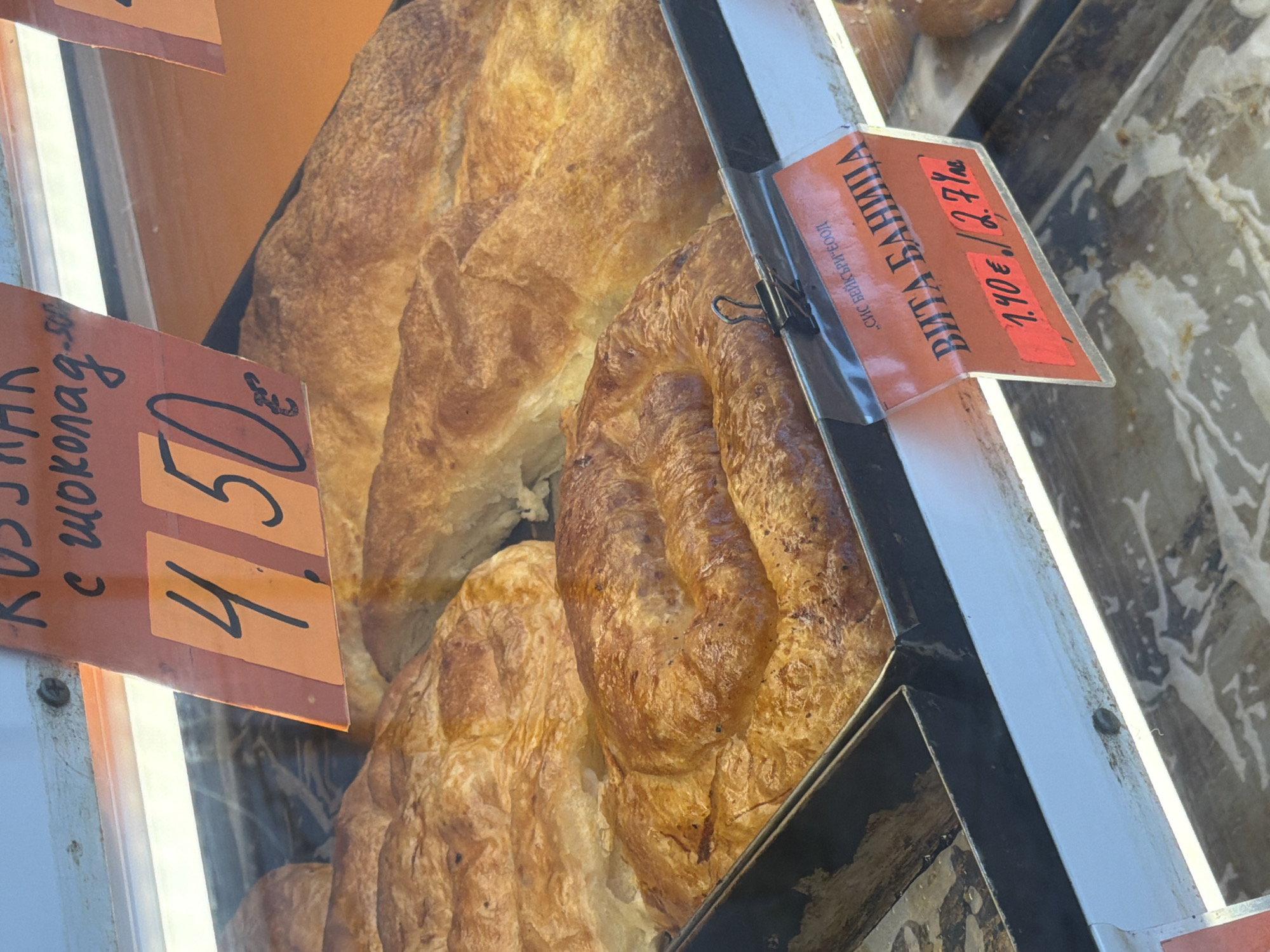 This image shows a display case of pastries in a bakery, likely located in Varna, Bulgaria. Two types of pastries are visible with their respective price tags. The pastry on the left is labeled "RUSJJA с шоколад" with a price of 4.50€. The pastry on the right is labeled "ВИТА БАНИЦА" and is priced at 1.40€/2.7лв per kilogram. A metal display case separates the pastries. The surface around the display case appears dirty.