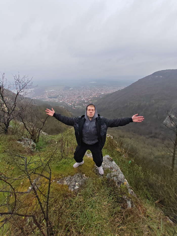 A young adult male stands centrally on a rocky outcrop, overlooking the city of Shumen, Bulgaria, situated in a valley below. He is wearing a dark grey hooded sweatshirt, an open black jacket, black pants, and light-colored athletic shoes. His arms are outstretched horizontally with open palms, and his eyes are closed. The immediate foreground features green grass and moss-covered rocks. To the left, sparse, bare-branched trees are visible. The midground consists of steep, forested hillsides, some trees still bare, others showing early foliage. The background reveals the urban sprawl of Shumen, characterized by dense residential areas with red-tiled roofs, extending into a distant haze. The sky is uniformly overcast and grey, providing diffused daylight.
