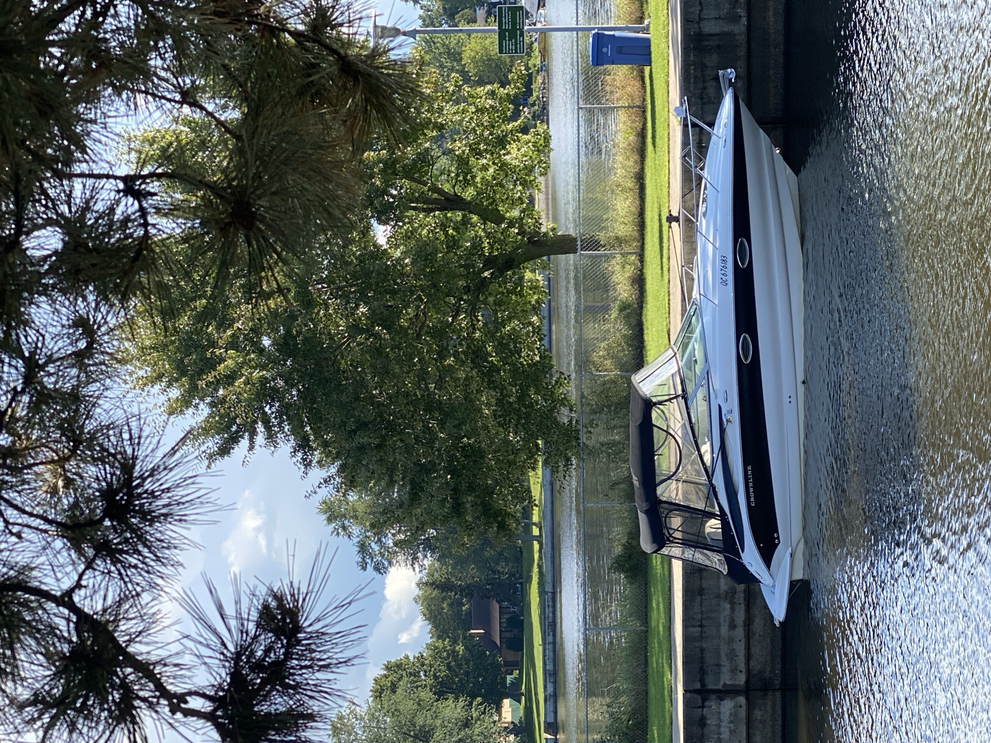 The image depicts a white and black Crownline boat docked along a concrete embankment on a calm body of water, likely a canal or river. The boat has a black canvas canopy extended over part of its cabin. In the background, across the water, is a grassy bank with a chain-link fence and mature trees. Beyond the trees, a residential house with a brown roof is visible. On the right side of the image, a blue garbage bin and a lamppost with a green informational sign are present. The scene is framed by the needles and branches of a coniferous tree in the foreground, suggesting an outdoor setting in Sainte-Anne-de-Bellevue, Canada, possibly within a park or waterfront area. The sky is partly cloudy.