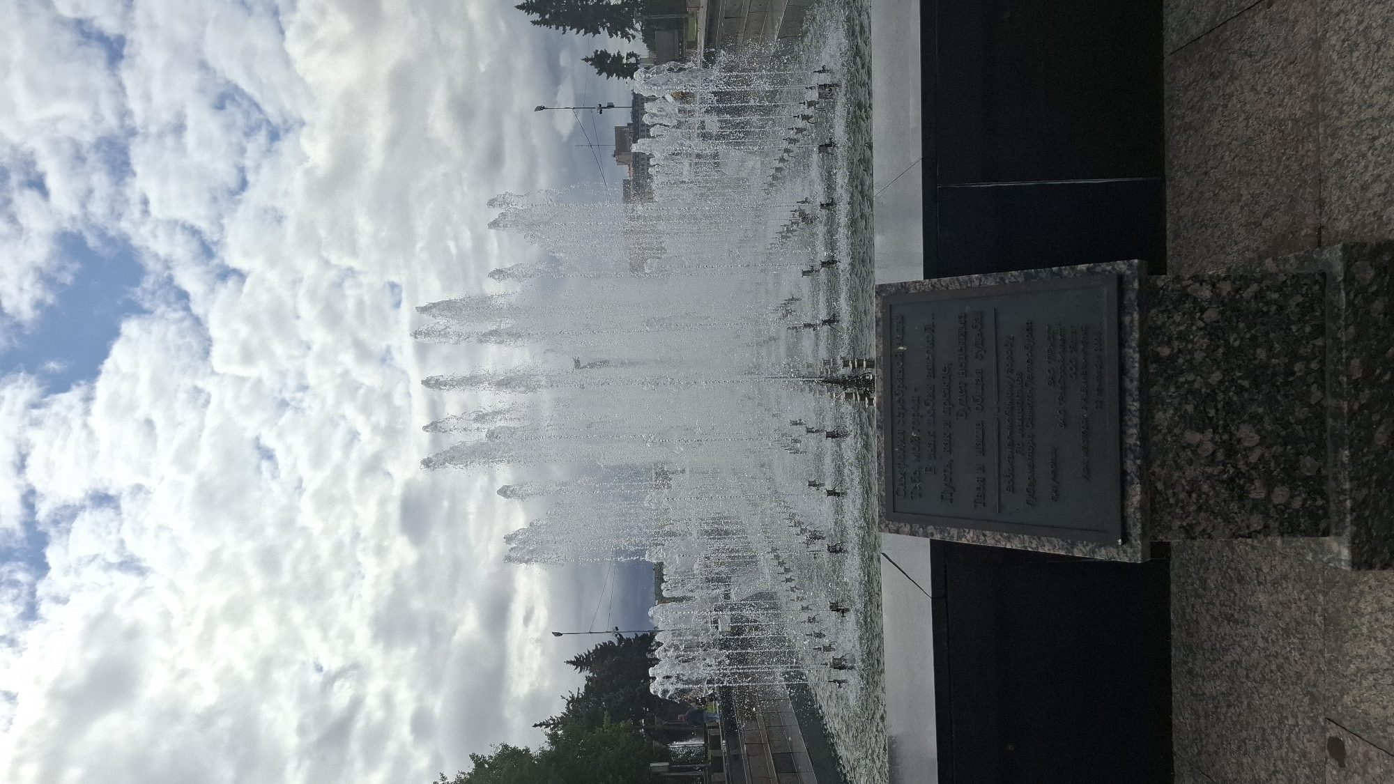 The image shows a large, multi-tiered fountain with numerous jets of water spraying upwards against a cloudy sky. In the foreground, a granite pedestal supports a dark plaque with an inscription. The fountain is situated in a plaza or park-like setting, with trees visible in the background. The inscription on the plaque appears to be in Russian and references "Saint Petersburg" and "Governor." Based on the inscription, this is likely a fountain dedicated to the city of Saint Petersburg, Russia. No people are prominently featured in the image, and the action is limited to the water from the fountain. The environment is an outdoor urban public space.