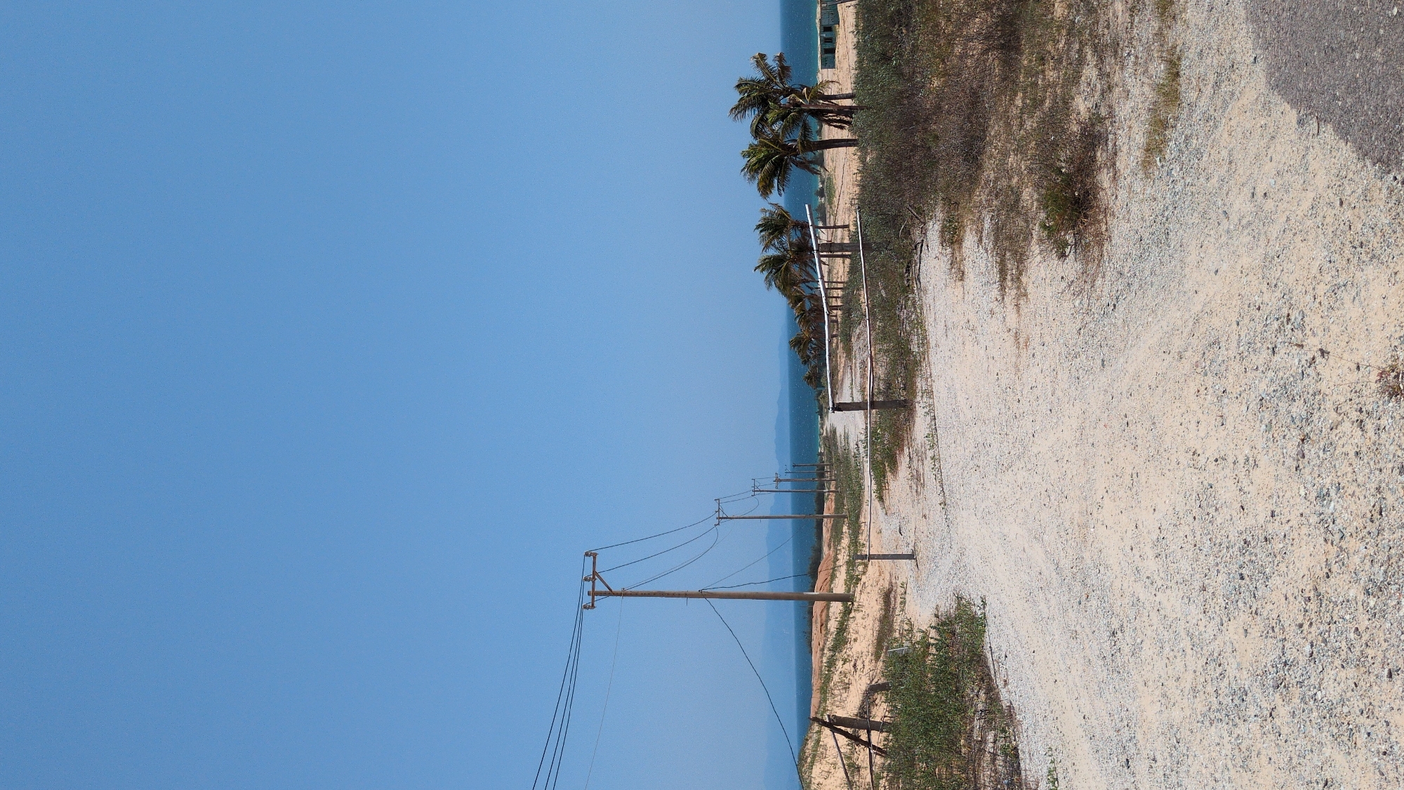 The image shows a landscape near the city of Phuoc Dinh, Vietnam, with a clear blue sky occupying the left side. A sandy or gravel path runs along the right side of the image. Several wooden utility poles with power lines stretch into the distance. Palm trees are visible along the shoreline. The ocean is present as a thin strip between the beach and the sky. Vegetation is scattered along the side of the path. A paved road is visible to the right of the gravel path.