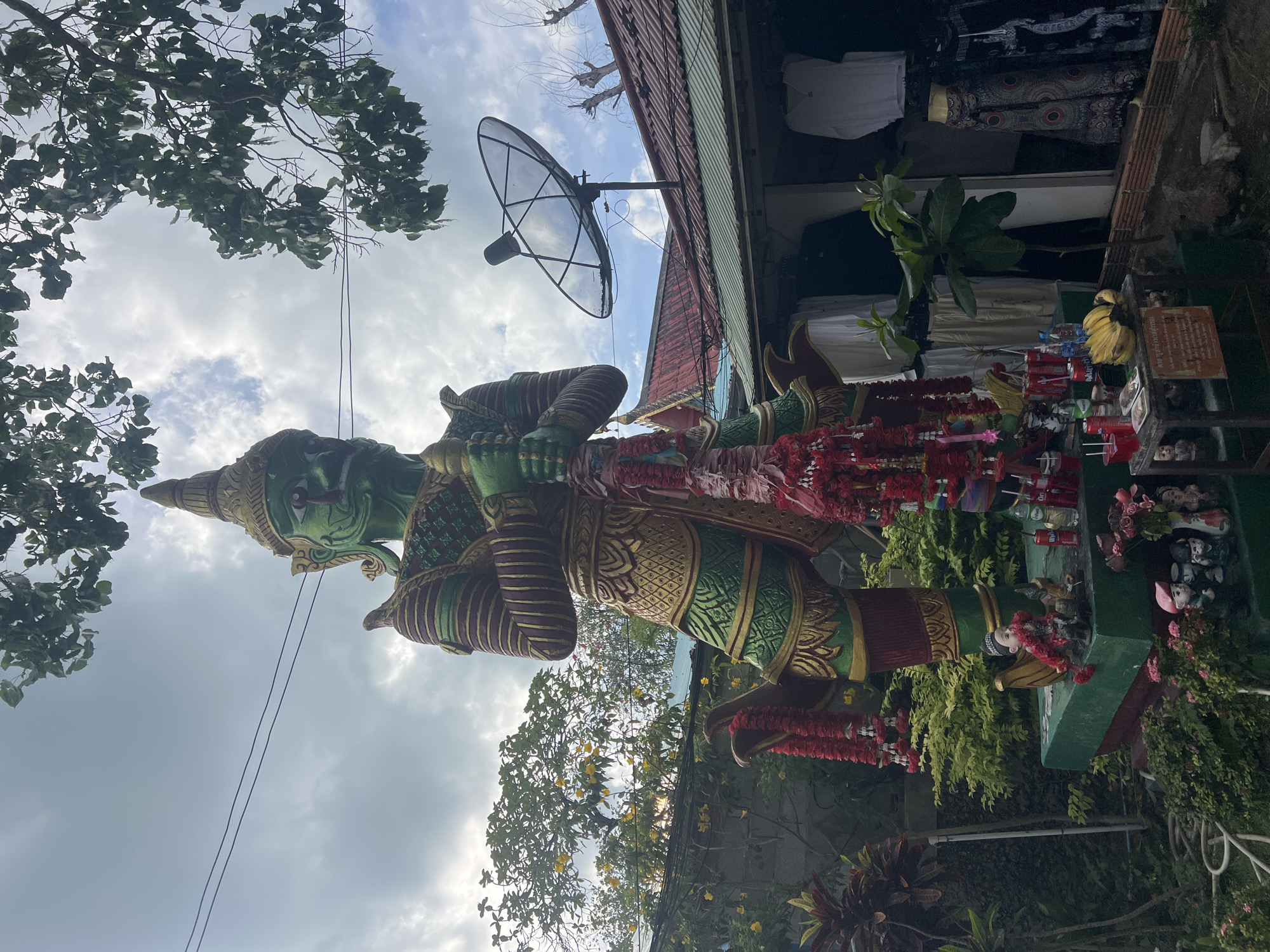 The image shows a large green statue of a mythical figure, possibly a Yaksha or similar guardian spirit, standing in what appears to be a public or semi-public space in Baan Plai Laem, Thailand. The statue is adorned with garlands of red flowers. To the right, there are offerings of bananas, drinks, and other items on a small altar-like structure. The background includes vegetation and the side of a building with a satellite dish. The sky is partly cloudy, indicating an outdoor setting during daylight hours. Clothes are hanging inside the roof of the building.