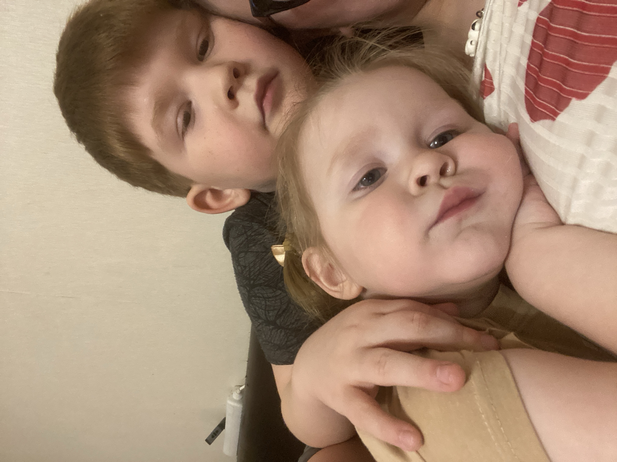 Two children are visible in a close-up indoor shot, with a partial view of an adult in the background. A boy with short brown hair wears a dark gray patterned shirt. A fair-skinned toddler with blonde hair pulled back in a small ponytail wears a tan-colored shirt. The adult in the background wears a white and red patterned shirt. The location is unknown, as it is not possible to determine it is in Chaikovsky, Russia. The children are looking towards the camera.