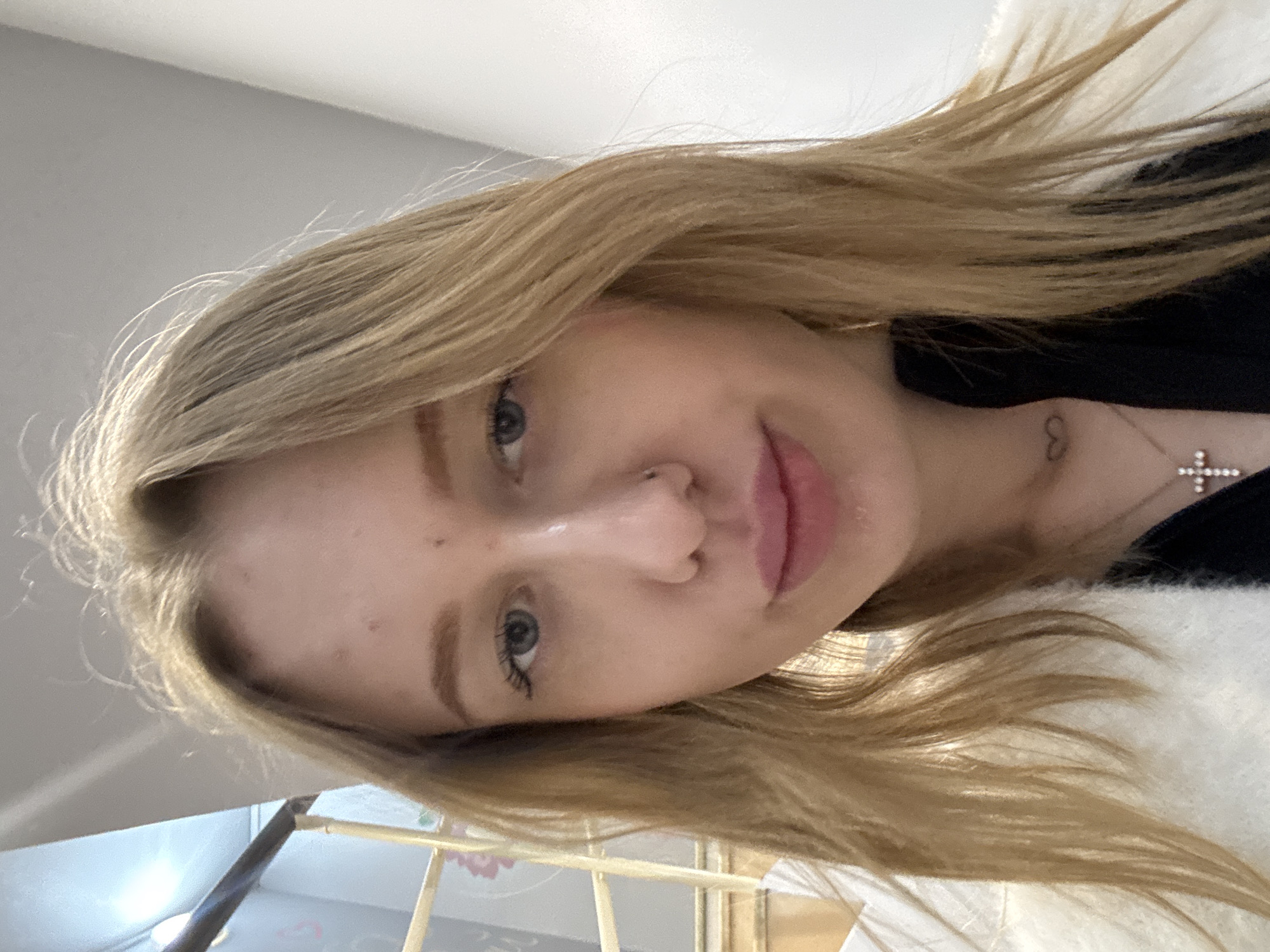 A close-up, rotated selfie of a young woman with light blonde, shoulder-length hair. She has fair skin, blue eyes, and pink lips. A silver cross necklace and a small tattoo (likely a heart shape) are visible on her neck. She is wearing a black top under a white sweater or jacket. The background is blurry and indistinct, with hints of a white wall and a metal frame, possibly a bed or furniture. There is insufficient information to determine the location in Rostov-on-Don.