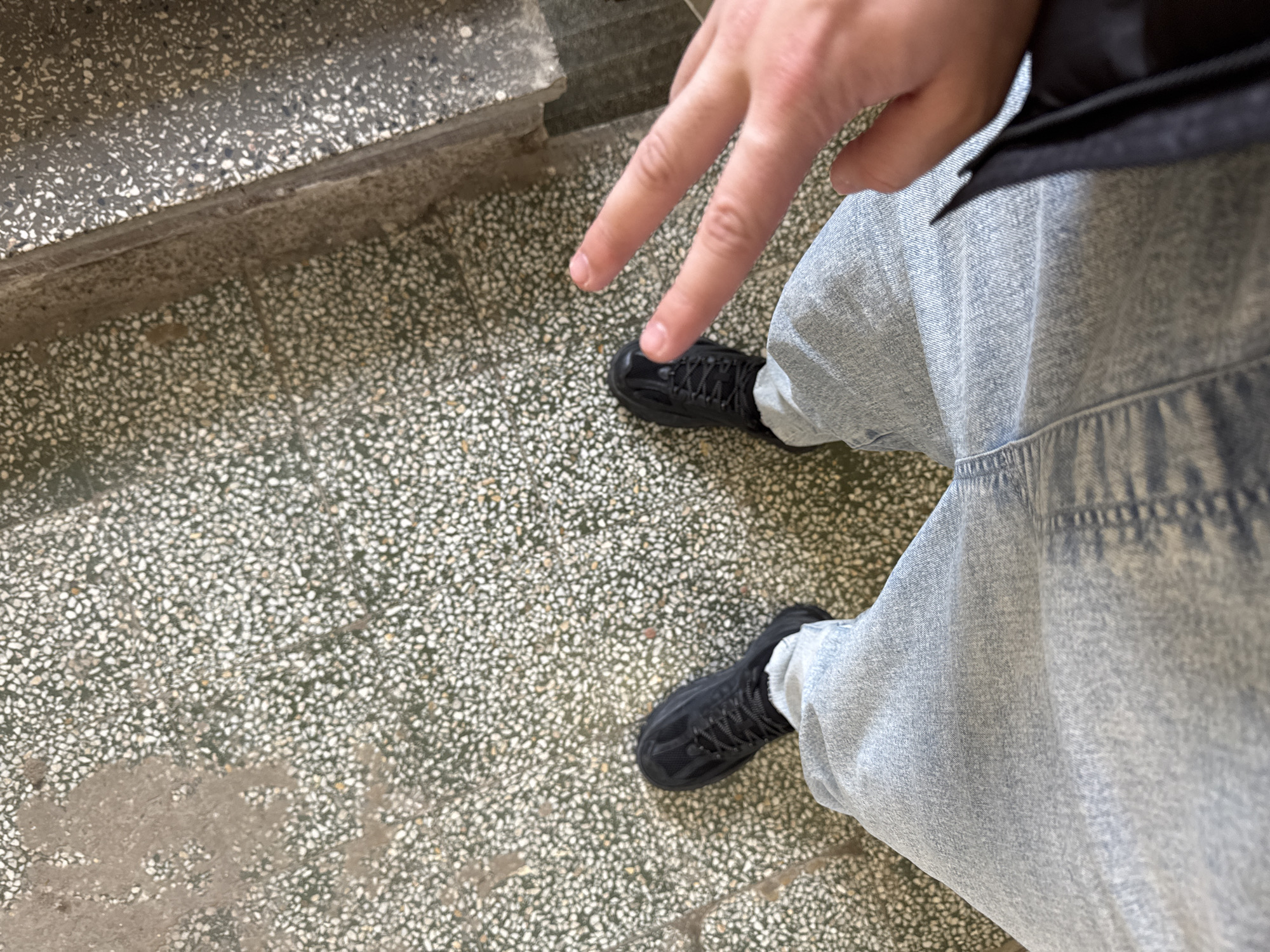The image depicts a person standing on a speckled, grey-and-white tiled surface. The person is wearing light blue denim pants and black sneakers. A hand with two fingers extended is visible above the left sneaker. The setting appears to be indoors, near a concrete step. The precise location in Sofia, Bulgaria is not discernible from the image.