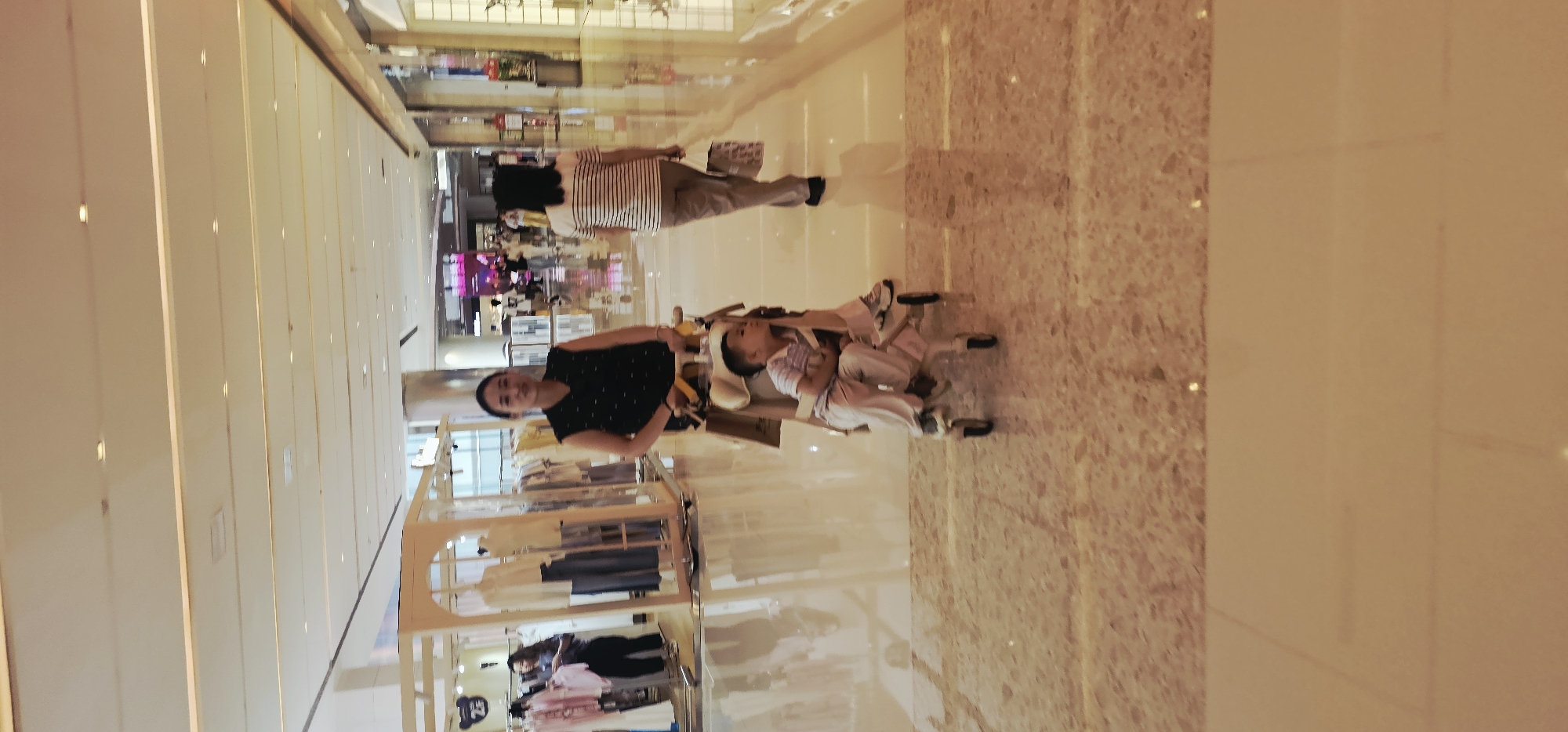 The image depicts a well-lit interior of a shopping mall, likely in Jiulongpo District, China, characterized by its polished floors and modern ceiling design with integrated lighting. In the foreground, a woman in a black sleeveless top with white polka dots pushes a stroller containing a young boy. The boy is wearing a striped shirt and light-colored pants. The woman is looking towards the camera with a neutral expression. Behind them, further down the mall corridor, another woman walks away from the camera, wearing a horizontally striped t-shirt and light brown pants, carrying a shopping bag. Storefronts are visible in the background, displaying clothing on mannequins and racks. The overall ambiance is one of everyday commercial activity.
