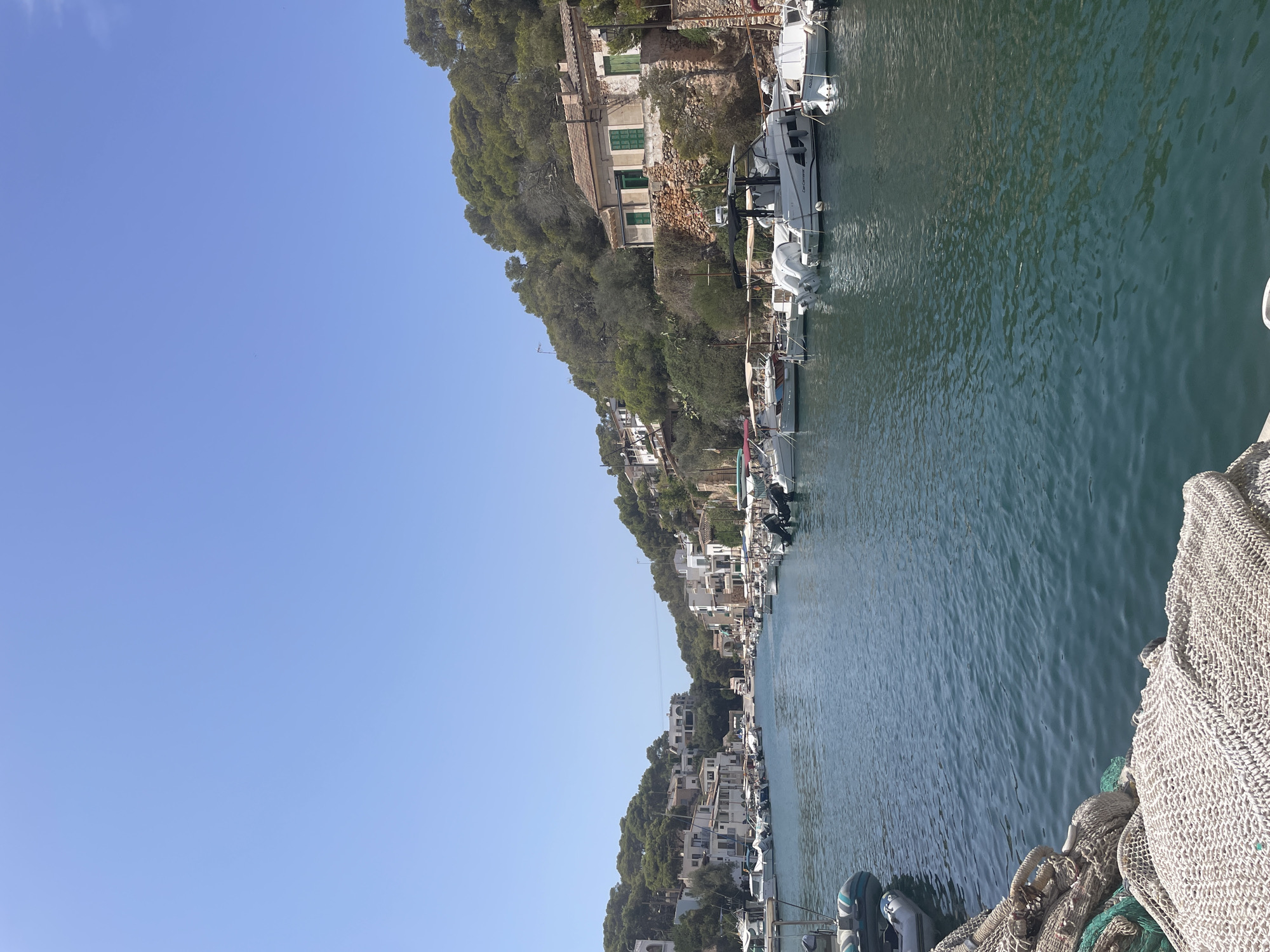 The image depicts a serene canal in Cala Figuera, a charming fishing village within the municipality of Santanyí, Spain. The canal is lined with traditional white houses adorned with green shutters, nestled into lush, tree-covered hillsides. Numerous small fishing boats and motorboats are moored along both sides of the canal, their reflections shimmering on the clear, blue water. In the foreground, fishing nets are piled on what appears to be a dock or embankment. The sky above is a vast expanse of clear, bright blue. The scene evokes a sense of peaceful, coastal life.