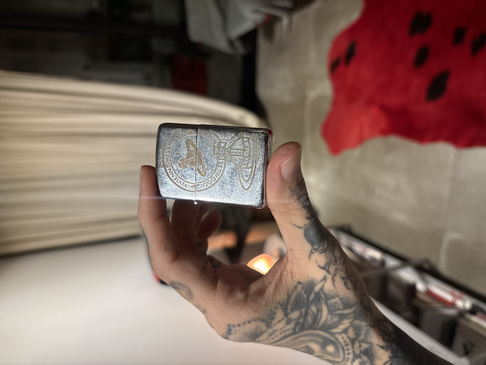 A person is holding a rectangular, silver-colored lighter in their right hand. The lighter has a design that includes the words "VIVIENNE WESTWOOD." The person's hand and lower arm are extensively tattooed with black ink designs. The background is blurred, but it appears to be an indoor setting. A red, polka-dotted object is visible in the upper right. The location is unknown and cannot be determined from this image. The city of Buenos Aires, Argentina, is not discernible from this image.