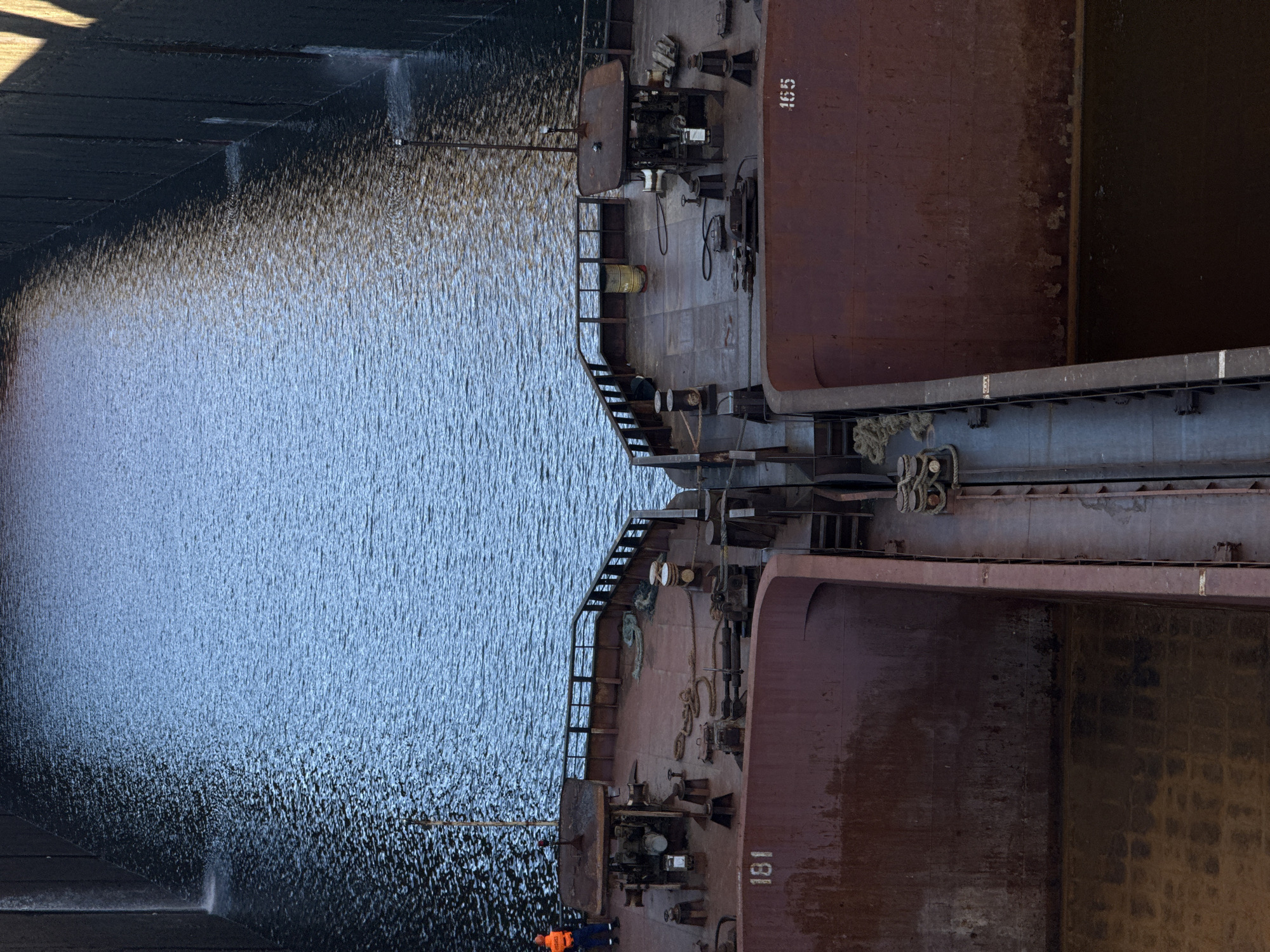 This media file shows a section of a lock chamber at a hydroelectric power station, likely the Votkinsk Dam in Chaikovsky, Russia. Two large, rusty barges are positioned within the chamber, which is filled with dark, rippling water. The water's surface is illuminated by sunlight, creating a shimmering effect.
On the deck of the upper barge, which has the number "165" stenciled on its side, various pieces of industrial equipment and machinery are visible, including winches, a large control panel, and piping. There are also ropes and metal railings. A single person wearing an orange vest is visible on the lower barge, near a set of stairs leading down to the water. This barge has the number "181" stenciled on its side.
The scene appears to be set during daylight, with clear weather indicated by the bright illumination of the water. The rough, weathered texture of the barges and the industrial setting suggest a functional and working environment. The presence of the barges within the lock chamber indicates an ongoing operation of navigating vessels through the dam, likely for cargo transport or maintenance. The overall impression is one of industrial activity within a waterway.