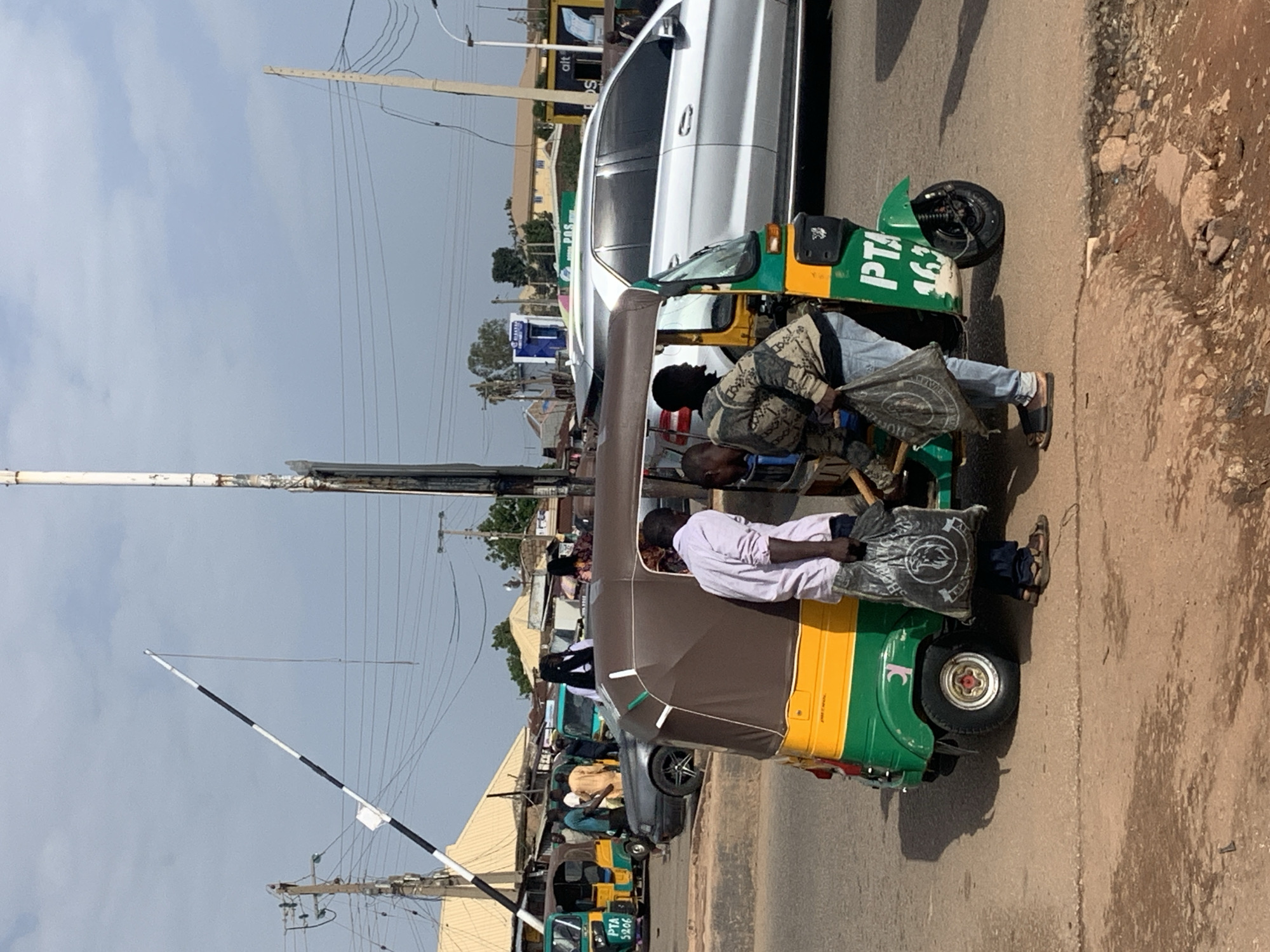 The image shows a street scene in Gura Top, Nigeria, featuring a green and yellow motorized rickshaw, also known as a "Keke Napep," parked on a dirt road. Two men are interacting with the rickshaw. The man on the left, wearing a light pink long-sleeved shirt and dark pants, holds a black plastic bag. The man on the right, dressed in a camouflage-patterned jacket and blue jeans, is also holding a similar black bag and appears to be placing it into the rickshaw or retrieving something from it.
In the background, other vehicles are visible, including a silver SUV and another Keke Napep. Several people can be seen in the vicinity, some standing and others near the vehicles. Buildings with yellow and white roofs are present in the background, suggesting a commercial or residential area. Overhead power lines and utility poles are also prominent features of the environment. A white and black striped barrier, possibly part of a checkpoint or toll booth, is positioned in the upper left portion of the frame. The sky is overcast with clouds. The overall scene depicts a moment of activity at a roadside location.
