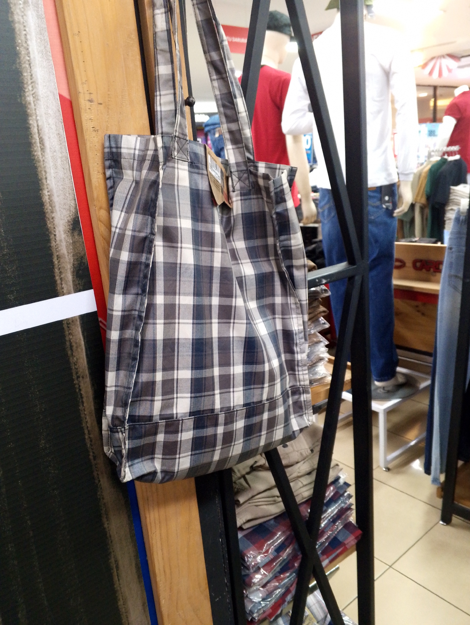 The image shows a close-up of a plaid tote bag hanging from a black metal rack in what appears to be a retail store, possibly a clothing store in Batam, Indonesia. The bag is primarily gray and white with blue and black accents in a checkered pattern. It has a simple design with two shoulder straps and a single compartment.
In the background, partially visible, are mannequins dressed in clothing, including a mannequin wearing a white long-sleeved shirt and blue jeans. Other clothing items, such as folded shirts and jeans, are also stacked on shelves or racks. The store environment has tiled flooring and displays of merchandise.
The lighting suggests an indoor setting, likely a commercial space. The overall impression is of a retail display focusing on apparel.