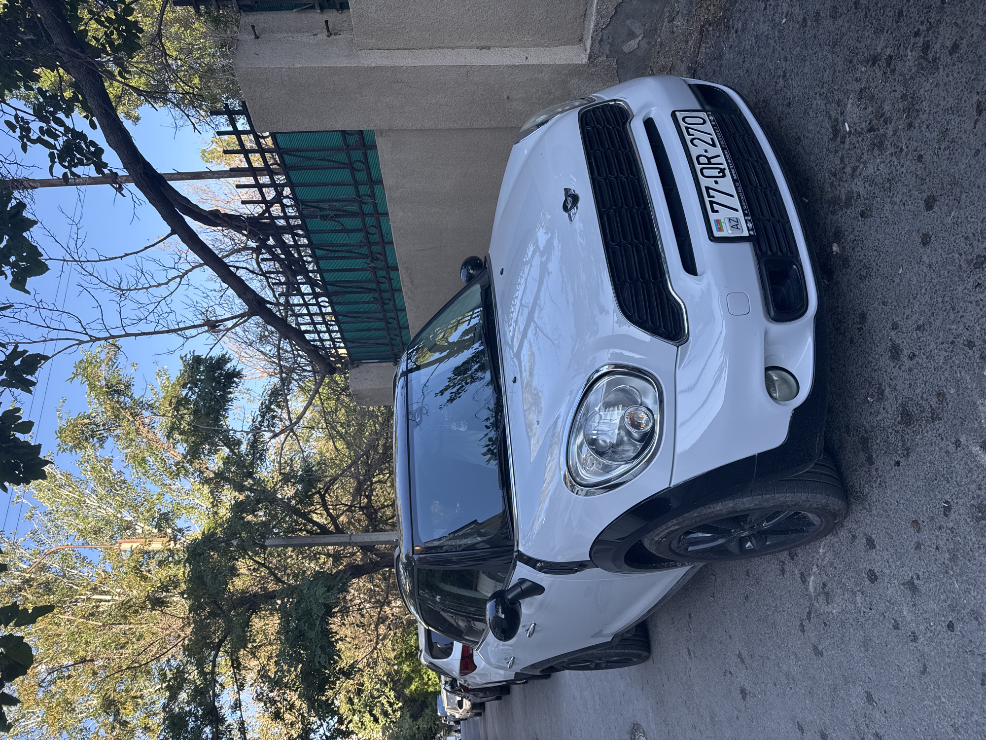 In Baku, Azerbaijan, a white Mini Countryman is parked on a paved street lined with trees and a decorative metal fence. The car, with its black roof and wheel arches, faces the viewer at a slight angle. Its license plate, "77-QR-270", is clearly visible. The street is partially shaded by trees, casting dappled light on the scene. The background shows a wall and more foliage, indicating a residential or urban setting. The sky is clear and blue. Several other cars are parked further down the street, suggesting a typical urban parking situation.