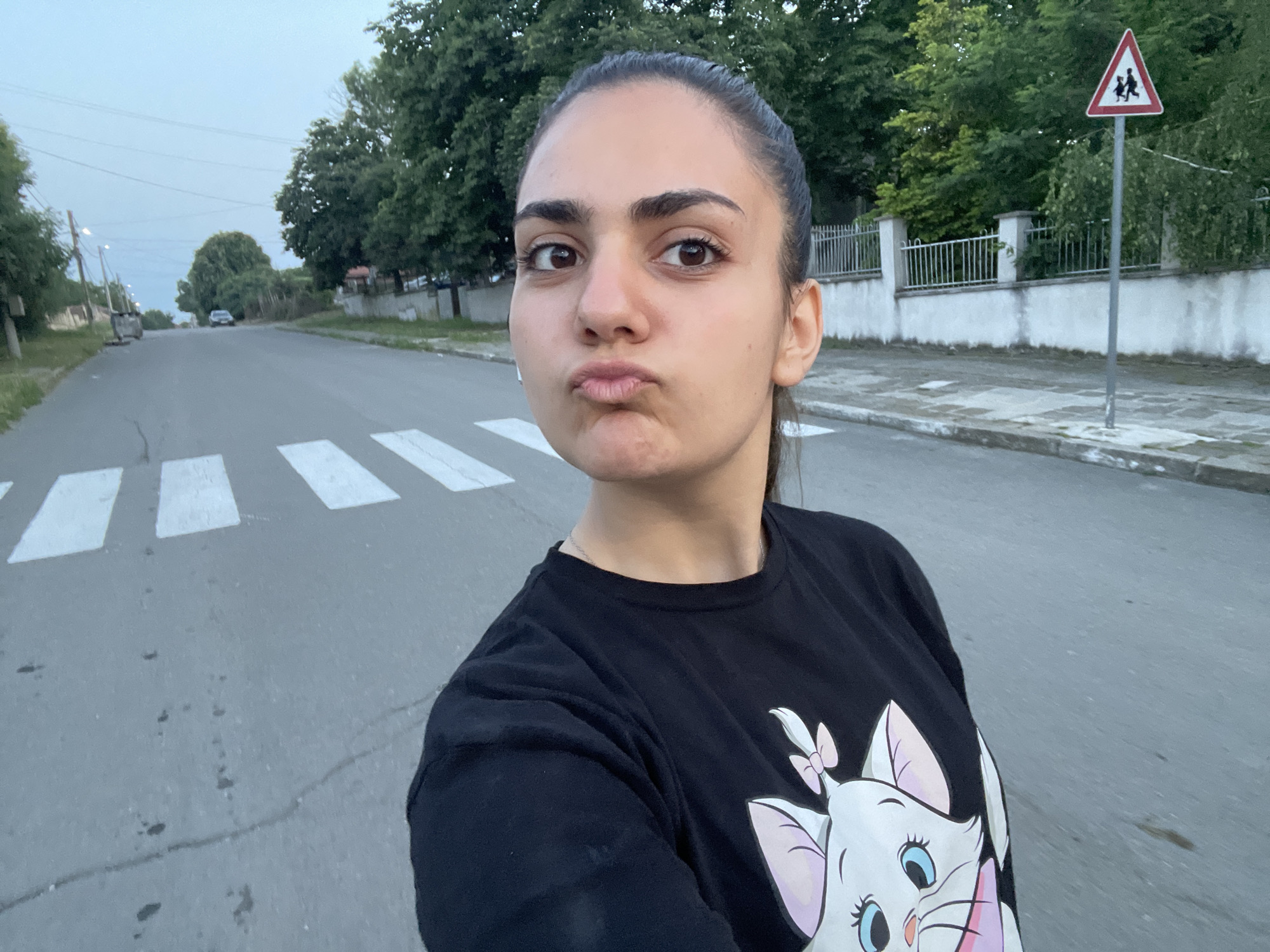 A young woman is shown in a self-portrait, positioned on a paved road marked with white pedestrian crossing stripes. She has dark hair styled in a ponytail and a pouting facial expression. She wears a black long-sleeved t-shirt featuring a graphic of Marie from Disney's *The Aristocats* on the front, along with a thin silver necklace. A white earbud is visible in her left ear. The asphalt road surface exhibits cracks and minor dark stains.
In the background on the left, the road extends into the distance, bordered by trees and streetlights, with a light-colored vehicle visible further down the street. On the right side of the image, behind the woman, a white concrete wall topped with a white metal picket fence runs parallel to the road. Behind the fence and wall, dense green trees and foliage are present. A red-bordered triangular road sign, depicting black silhouettes of an adult and a child, is mounted on a gray pole situated on the right side of the road. The sky above is overcast and light blue. The scene is located in Debelt, Bulgaria.