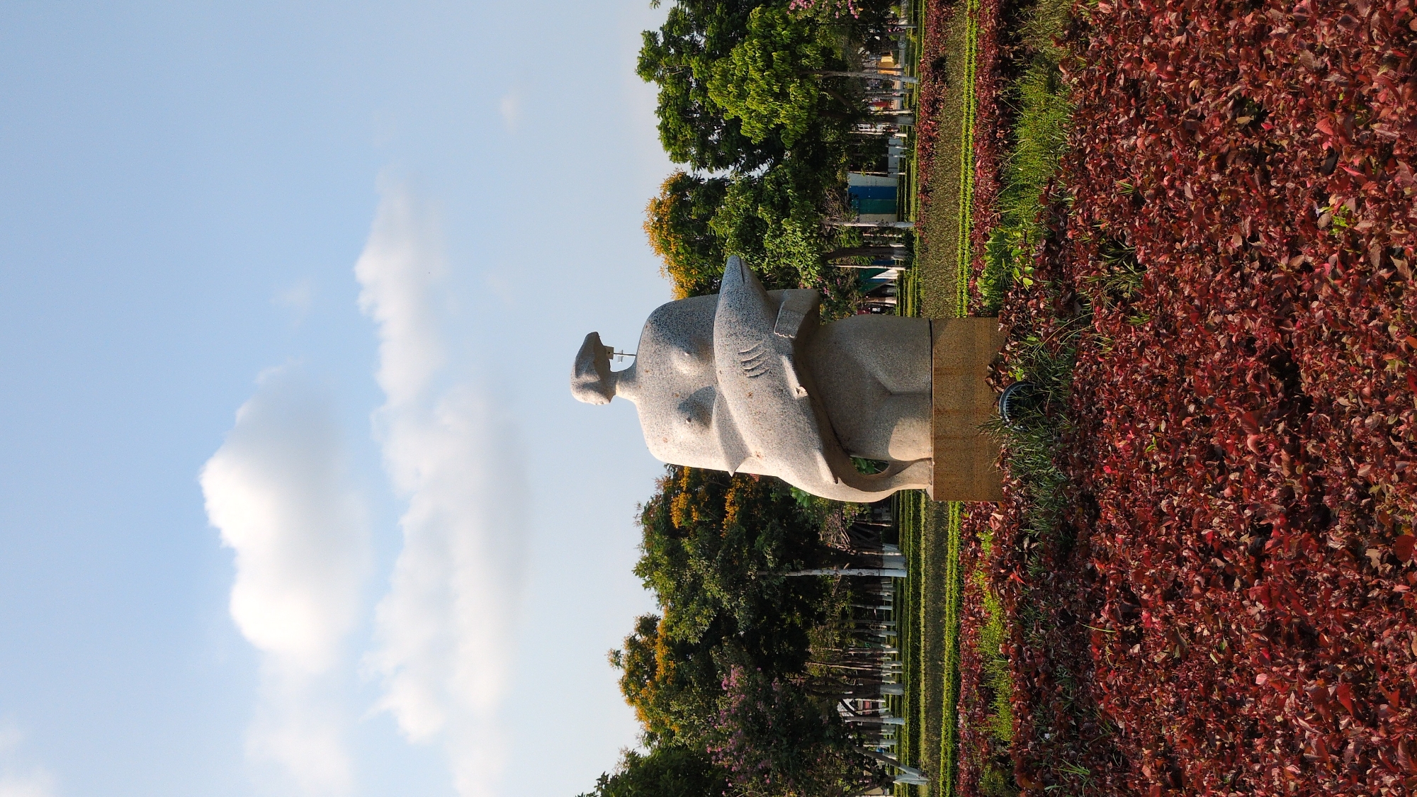 A statue of a nude woman embracing a fish stands in a park in Phan Rang – Tháp Chàm, Vietnam. The statue is made of light-colored stone and is positioned in a bed of red plants. Behind the statue, there are trees, a green strip of lawn, and metal barriers. The sky is visible in the background, with a few clouds.