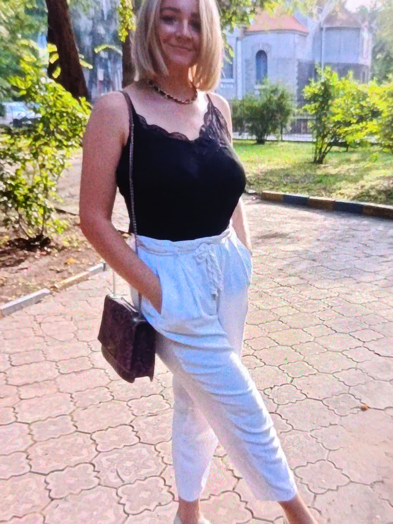 A woman with blonde hair is standing outside on a paved walkway. She is wearing a black lace-trimmed tank top and white pants. She has a dark-colored handbag with a chain strap slung over her shoulder. Her right hand is in her pocket.
The setting appears to be a park or a public area with trees and greenery in the background. There are buildings visible behind the trees. The paving stones on the walkway are a light color with a decorative pattern. A curb separates the walkway from a grassy area. The lighting suggests it is daytime, possibly late afternoon, with warm sunlight. The woman is smiling, conveying a sense of happiness or contentment.
There is no visible text in the image. The location context provided is Bălți, Moldova.
