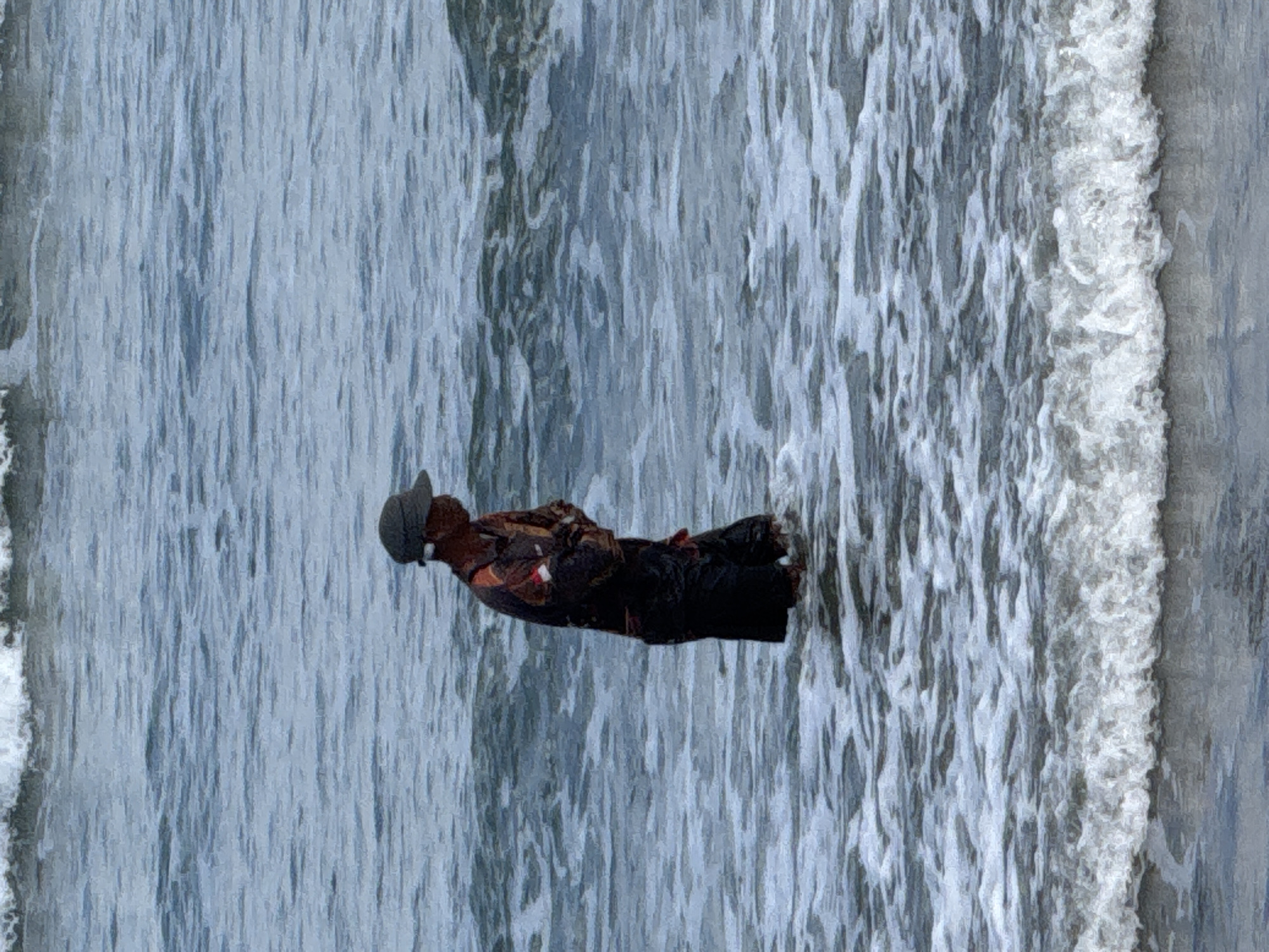 The image shows a person standing in the shallow water at the edge of the ocean in Kuta, Indonesia. The person is wearing a dark-colored shirt with orange and white accents, dark pants, and a dark cap. They are facing to the right and appear to be looking down. The water is choppy with white foam from small waves breaking around their legs. The background is filled with the blue-grey expanse of the ocean, with larger waves breaking further out. The lighting suggests it is daytime, possibly overcast given the diffused light.