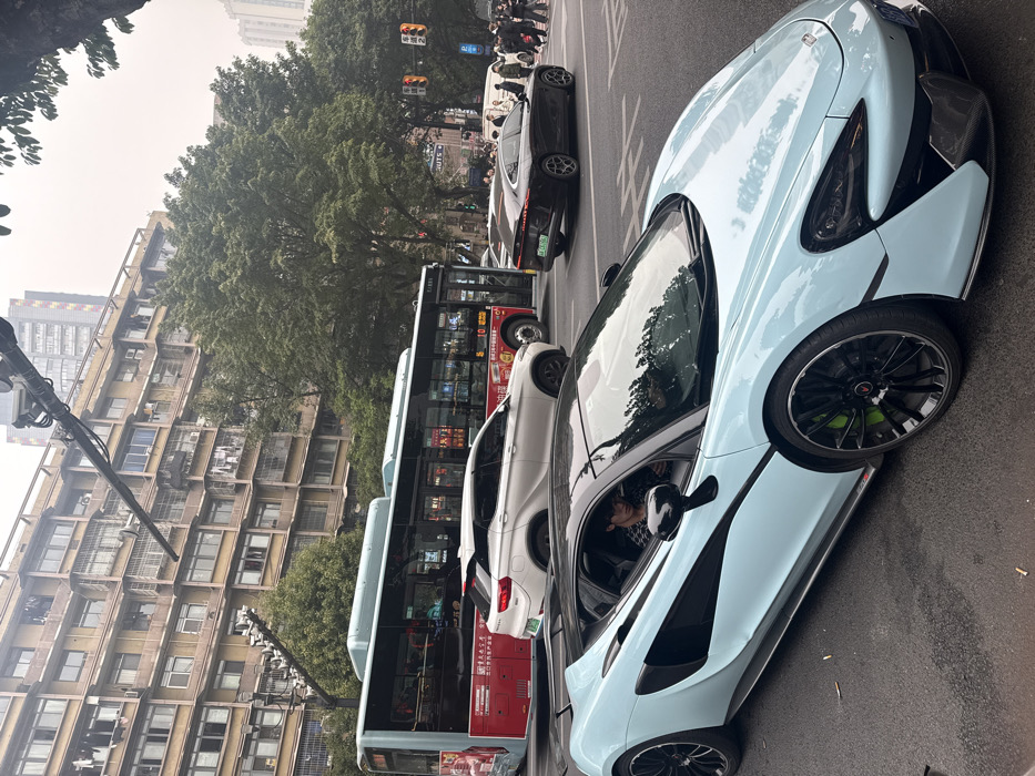 A light blue McLaren supercar is parked on the right side of a street, facing forward. To its left are a white SUV, a red and blue city bus, and a dark-colored sedan, all lined up in traffic. In the background, an older apartment building is partially visible on the left, with green trees and a traffic light in the distance. The overall scene suggests a busy urban environment.