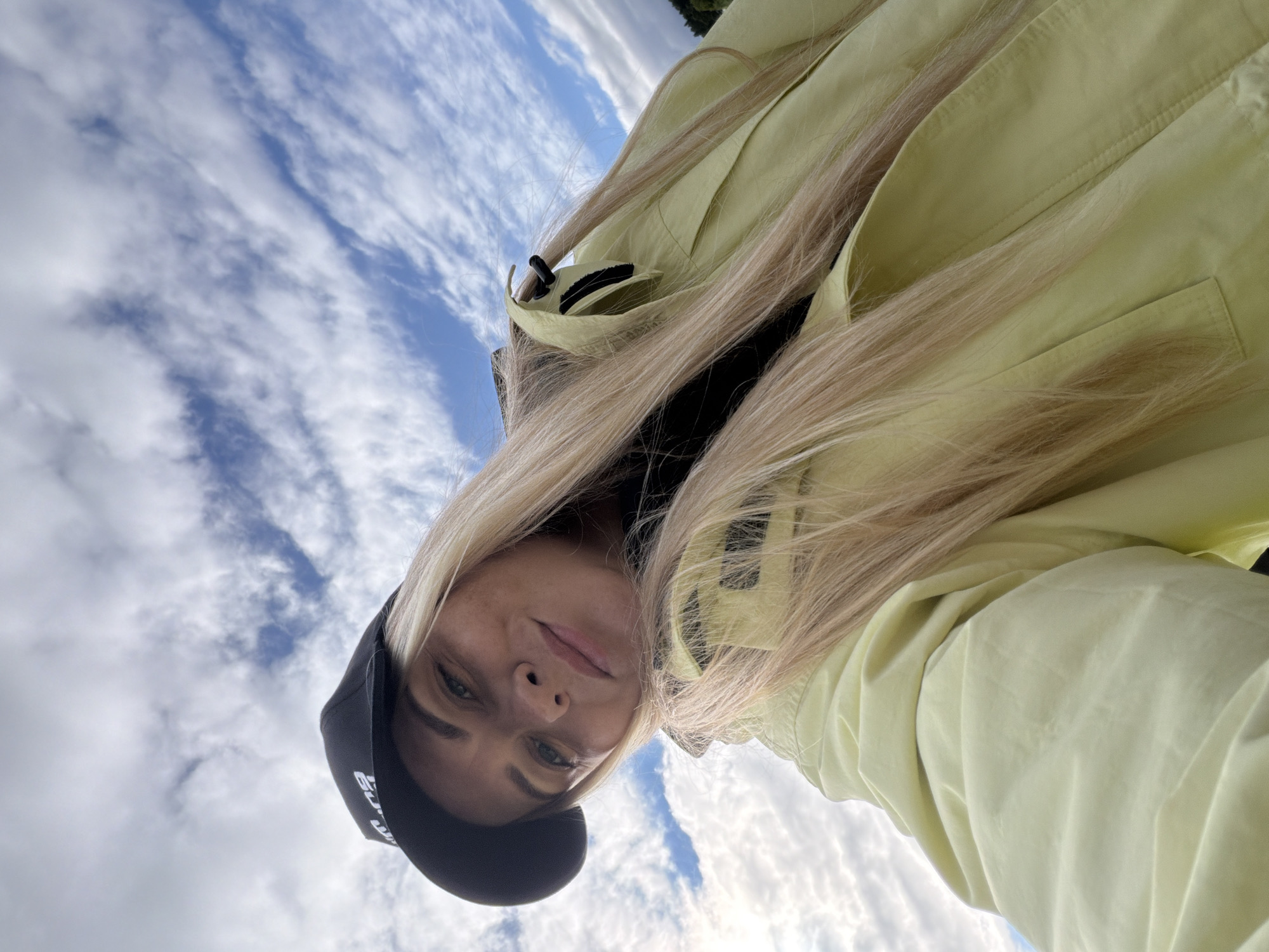 The image shows a close-up, selfie-style shot of a young woman outdoors, looking up towards the sky. She has long, blonde hair, is wearing a black baseball cap, and a light yellow jacket. The background consists of a bright blue sky with scattered white clouds. There is no visible indication of the city of Votkinsk, Russia, or any specific urban environment. The focus is on the woman and the sky.