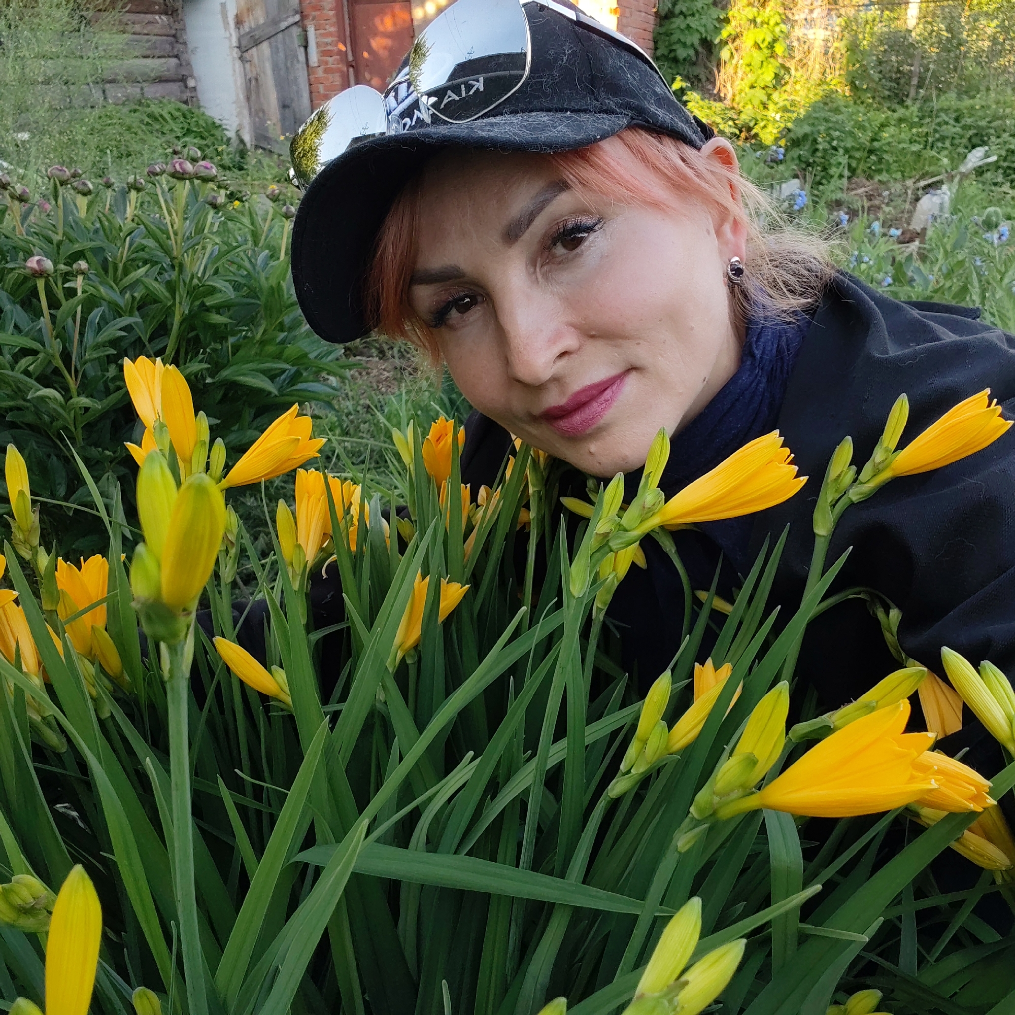 A woman with reddish-orange hair is captured in a close-up portrait amidst a lush garden, likely in Chaikovsky, Russia. She wears a black baseball cap with silver-mirrored sunglasses resting on top, which reflect what appears to be the word "KIVAIK". Her attire includes a black jacket and a dark blue top or scarf. She has visible makeup, including eyeliner, mascara, and pink lipstick, and looks directly at the camera with a subtle smile, conveying a calm and friendly demeanor.
The foreground and midground are dominated by numerous vibrant yellow daylilies (Hemerocallis), many still in bud, surrounded by their long, green foliage. The woman's upper body is positioned within or closely behind these flowers.
The background reveals a rustic outdoor setting, featuring parts of an old wooden building or structure, possibly a shed or house, and reddish brickwork. Green foliage and trees are also prominent, with small blue flowers scattered among the distant greenery. The warm, soft lighting suggests the photograph was taken during the late afternoon or early evening.