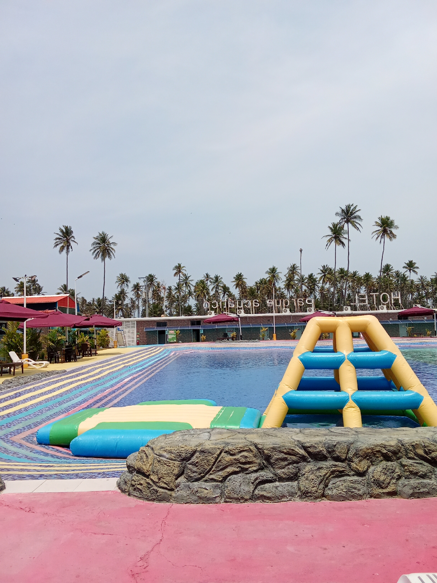The image depicts an outdoor water park at a hotel in Las Delicias, Venezuela. The water park features a large swimming pool with an inflatable obstacle course. The obstacle course includes a yellow and blue inflatable climbing structure and a green and blue inflatable floating platform. A faux rock wall is in the foreground. The pool deck is painted with a pattern of rainbow-colored stripes. In the background, there are palm trees, red umbrellas, and outdoor seating. A sign reads "HOTEL" in reverse. The sky is overcast. There are no people visible in the image.