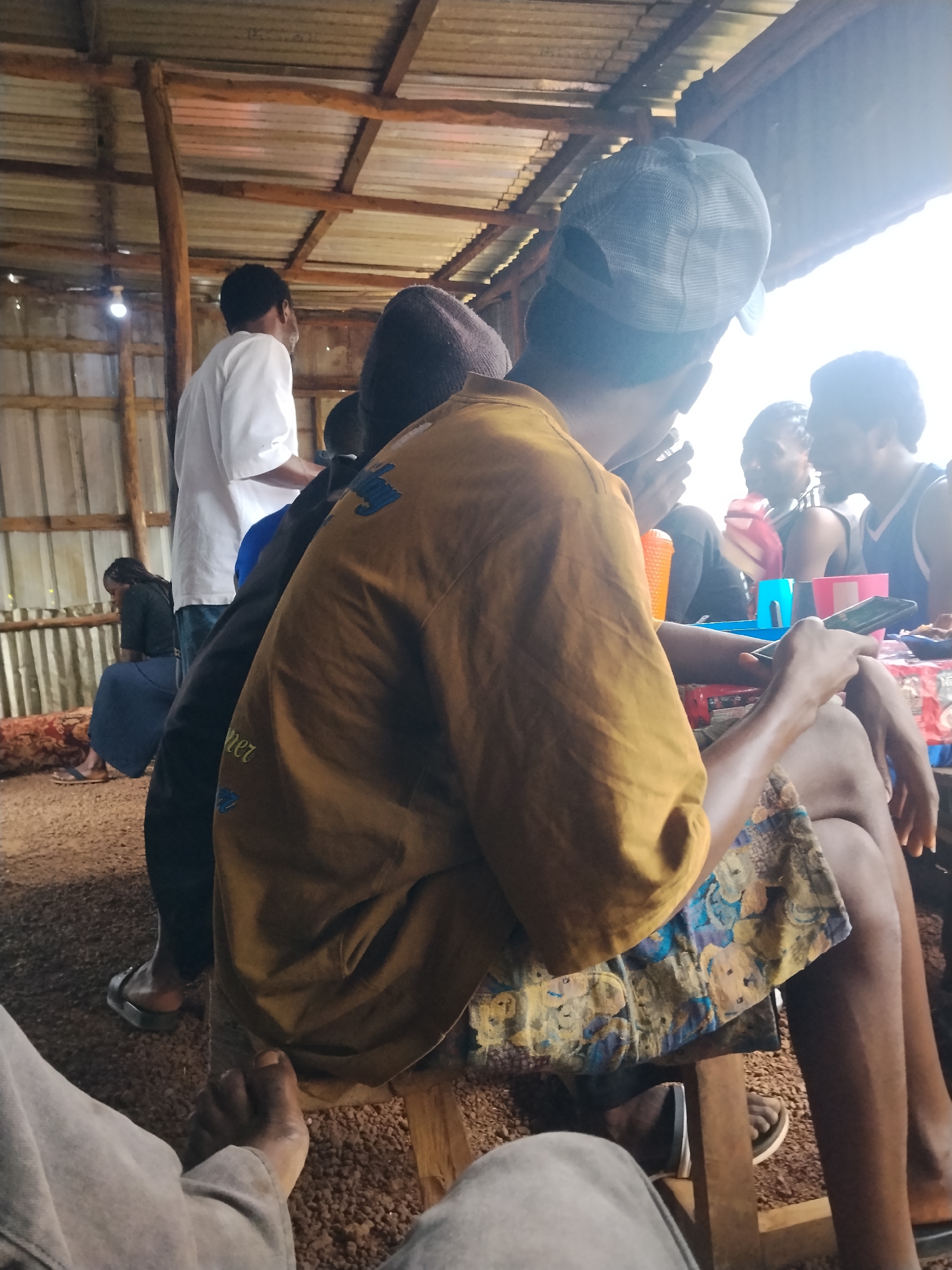 In Durumi, Nigeria, a group of people are gathered indoors at what appears to be a makeshift shelter with a corrugated metal roof supported by wooden beams. In the foreground, a person is seated, holding a mobile phone and wearing a yellow t-shirt and patterned shorts. To the left, a person in a white shirt stands facing away from the camera, while further left, another individual sits, facing away and wearing a dark top and a blue skirt. In the background, several other individuals are visible, some interacting with each other. One person wears a light-colored baseball cap. Various objects, including plastic cups and what might be plates of food, are present on a surface in front of the seated individuals. The ground is covered with small stones or gravel. The overall atmosphere suggests a casual gathering.