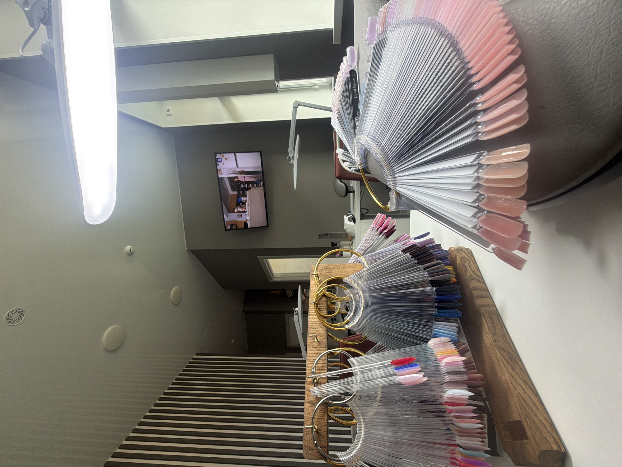 This image captures the interior of what appears to be a modern nail salon or beauty studio, shot with a strong diagonal tilt. In the foreground and midground on the right, multiple fan-shaped displays showcase a vast array of nail polish color swatches, organized on a wooden stand with prominent gold and silver rings. The colors span a wide spectrum, including numerous shades of pink, nude, brown, red, blue, and other vibrant hues. Visible text on one swatch reads "Liquid PolyGel L3" and "BASE L RD 5".
On the left side of the frame, a long, bright LED light fixture provides dominant illumination. Below it, a distinctive staircase or ramp descends, featuring alternating black and light-colored stripes. The adjacent light-colored wall displays circular vents or speakers and a smoke detector. In the background, a flat-screen television mounted on the wall shows a video with blurred figures, and a flexible-arm lamp is positioned nearby. No individuals are directly visible in the scene, but the presence of nail polish swatches clearly indicates a setting for nail services. The overall atmosphere is well-lit, organized, and functional. The specific location is Izhevsk, Russia.
