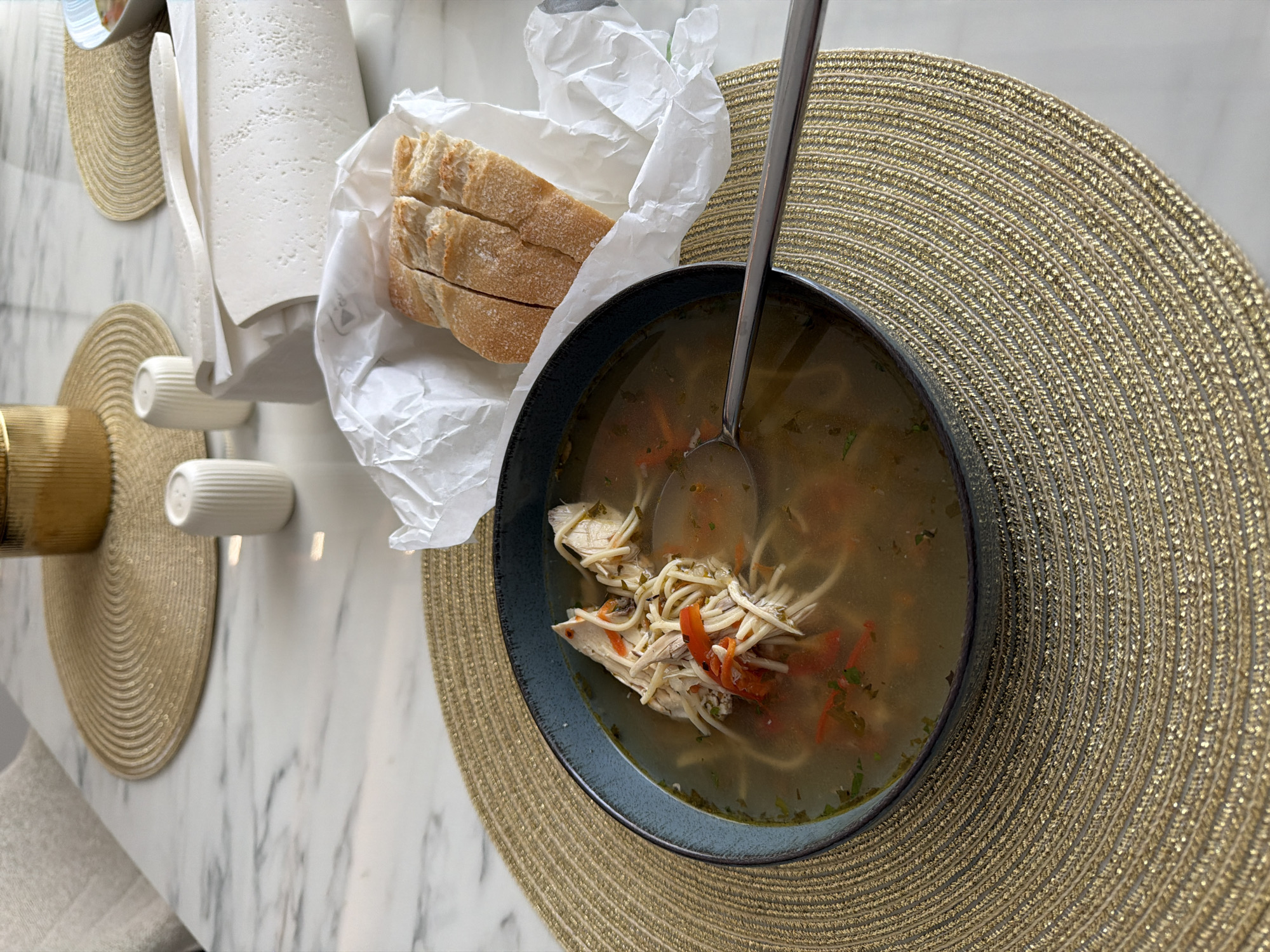 A close-up, eye-level shot shows a rustic dark blue bowl filled with chicken noodle soup. The soup contains shredded chicken, thin noodles, and chopped red peppers in a clear broth, with a silver spoon resting in the bowl. The bowl is positioned on a circular, textured gold placemat. To the right of the soup, a loaf of sliced baguette is presented in white parchment paper. In the background, a white marble table is visible, with a gold cylindrical object, two white salt and pepper shakers, and a large block of white cheese wrapped in white paper. The setting appears to be an indoor dining area in Chișinău, Moldova, possibly a home or restaurant, suggesting a meal is about to be or is being consumed.