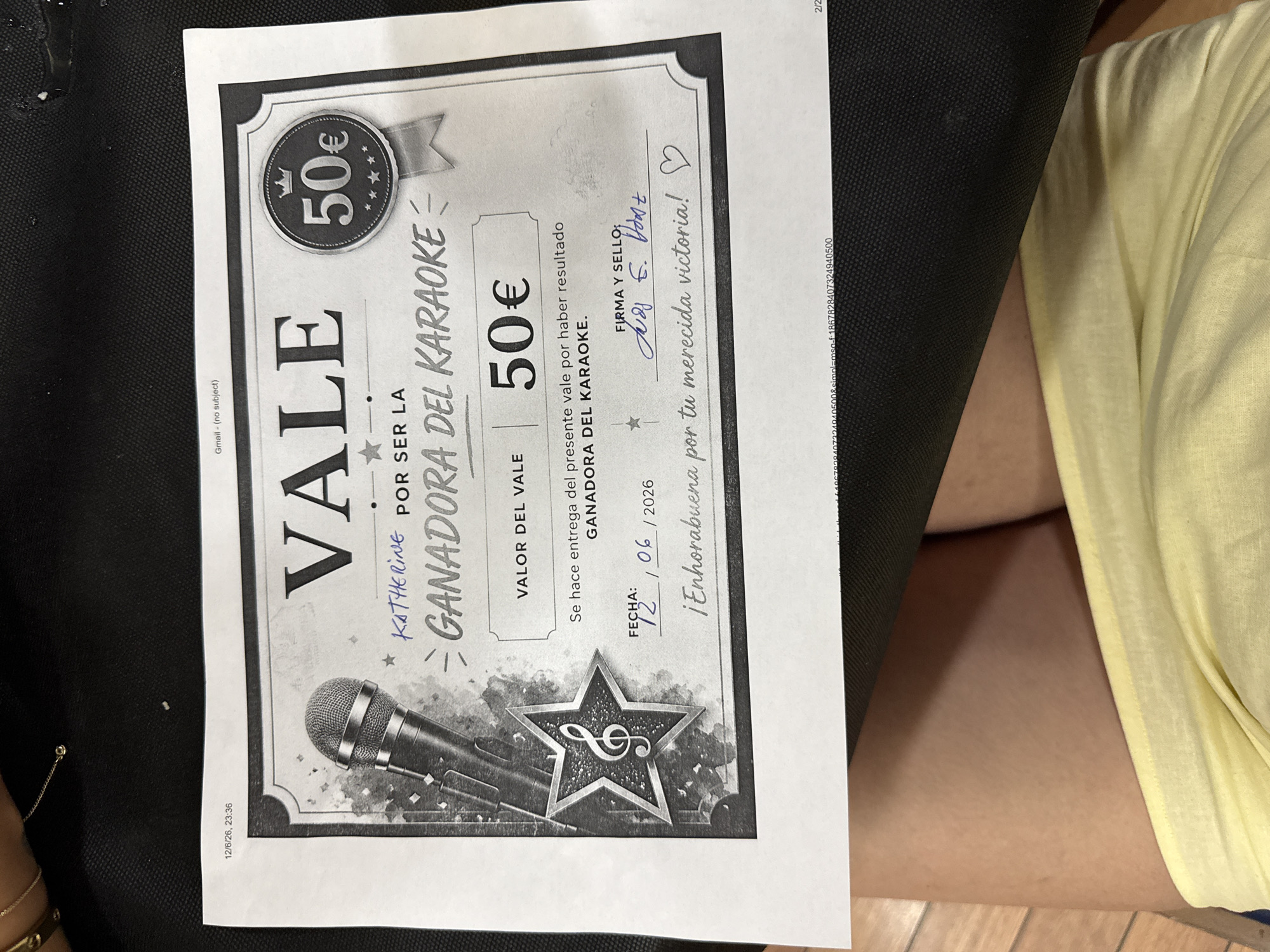 The image shows a "VALE" (voucher) for "GANADORA DEL KARAOKE" (Karaoke Winner). The voucher is for 50€. The date is written as 12/06/2026 and the signature and seal area has handwritten text. The voucher is printed on paper and is the central subject of the image. The voucher is placed on a dark textured surface, possibly a bag or a mat. A small portion of a person's legs wearing yellow clothing is visible at the bottom of the image. The lighting suggests an indoor setting. No specific location in Torrejón de Ardoz, Spain, can be determined from the image.