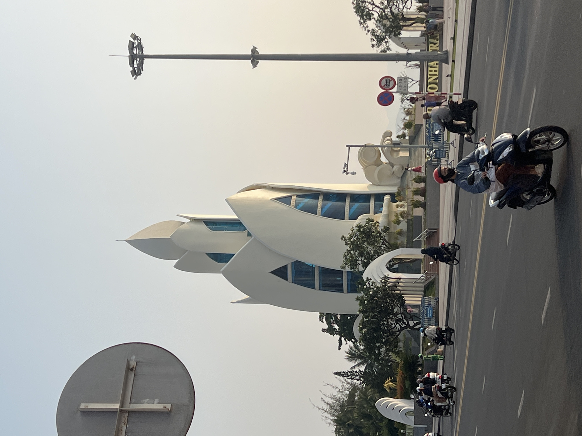 The image captures a street scene in Nha Trang, Vietnam. A modern, white architectural structure with a blue-windowed facade is the central element. Street signage, including road markings and a tall light pole, are present. Motorbikes are in motion on the road. The sky is overcast. Trees and other vegetation are visible around the building and along the road.