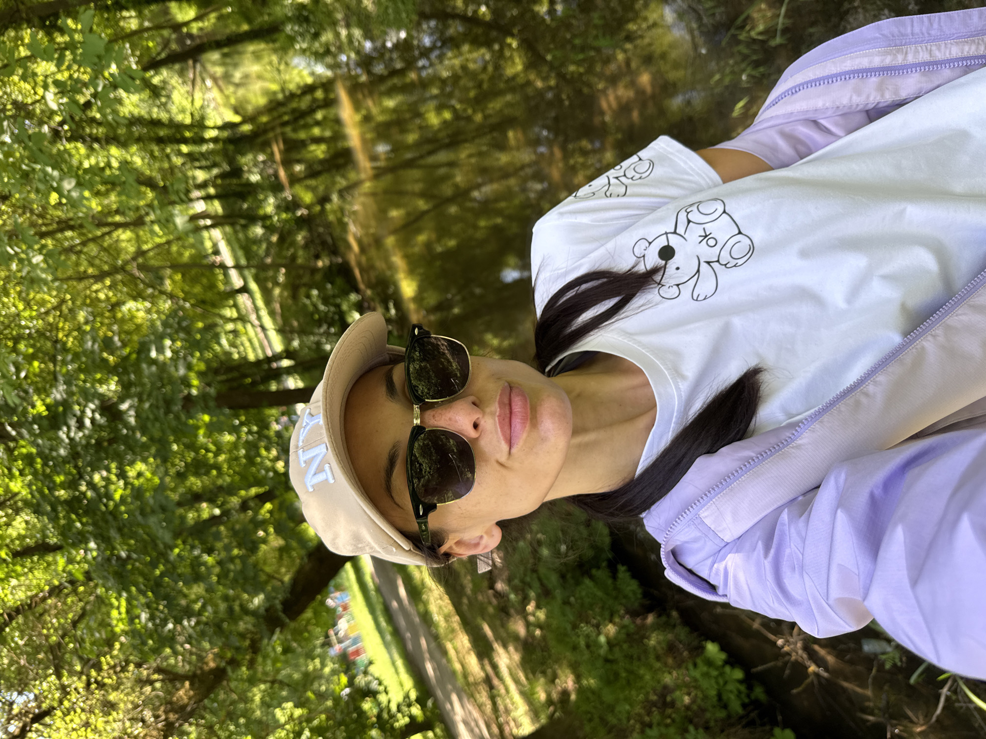 A young woman wearing a light beige "NY" baseball cap, dark sunglasses, a white t-shirt with bear graphics, and a light purple jacket is the main subject. She is outdoors in a park-like setting with abundant greenery. Behind her is a body of water reflecting the trees. The location is likely in Pleven, Bulgaria.