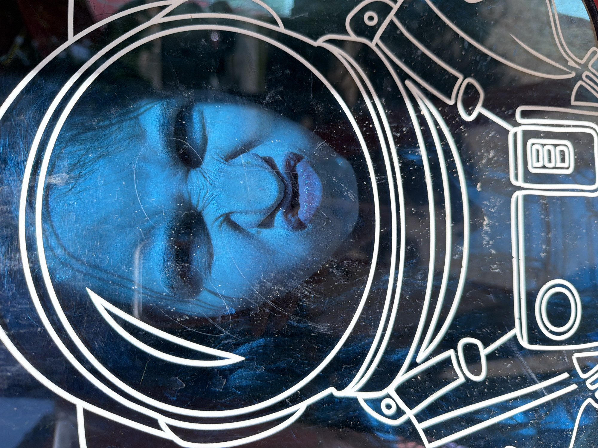 The image shows a person's face reflected in a display window, the reflection is distorted and blue-toned. The window is decorated with a white design, possibly a stylized drawing of a spaceship or astronaut's helmet. Based on available information, the photo was taken in Yerevan, Armenia.