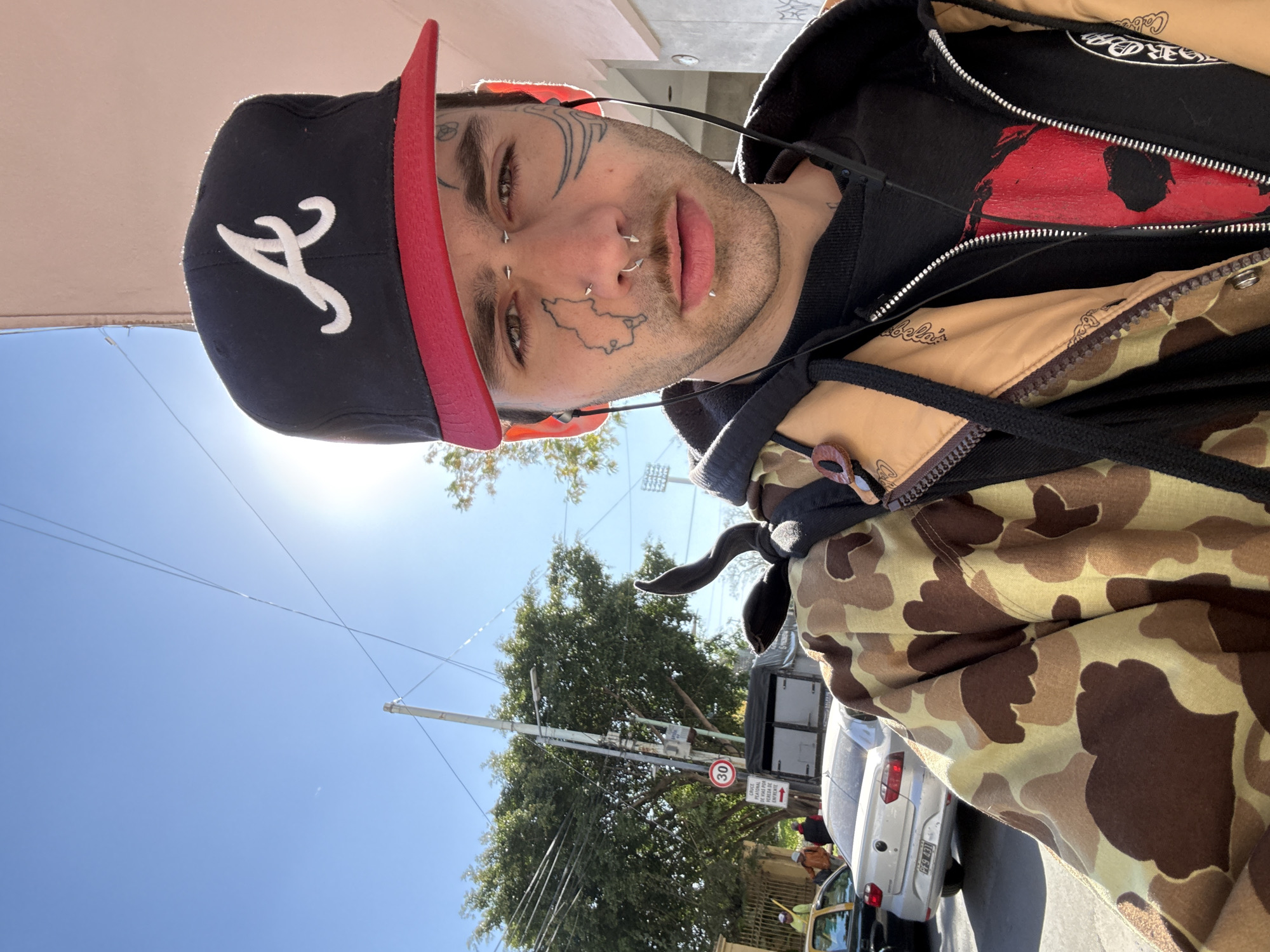 A close-up, low-angle shot features a person wearing a black baseball cap with a red brim and a white emblem on the front. The individual has multiple facial tattoos and piercings, wearing a black hoodie under a Cabela's brand camo jacket. The person is wearing earbuds with the wire visible.
In the background, the environment indicates an urban setting in Buenos Aires, Argentina. Street signs are visible, including a speed limit sign of 30 km/h. A line of parked cars lines the curb, including a white sedan with a license plate, partially visible as "925 481." People are also visible in the background near the vehicles. Overhead, the clear sky features a bright sun. A utility pole and power lines are also captured within the frame. The picture appears to be a selfie.