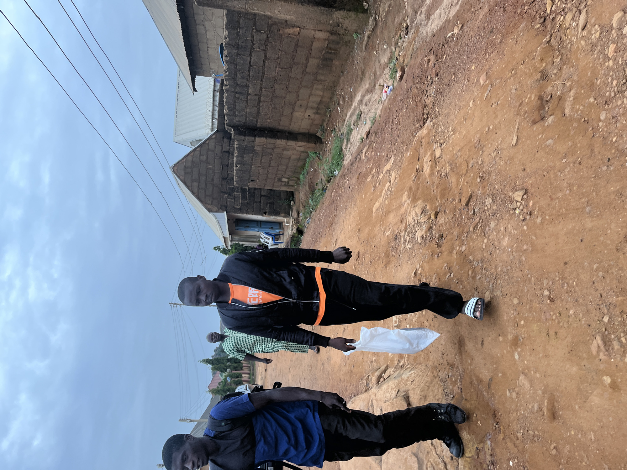 In the city of Jos, Nigeria, under a cloudy sky, three individuals are walking along a dirt road. The man in the foreground on the right is wearing a black jacket over an orange t-shirt and black pants, carrying a white plastic bag. To his left, another man is walking in a blue and black top and black pants, with a backpack. Behind them, a third man is walking in a green and white patterned robe. The road is unpaved and reddish-brown in color, with scattered rocks and debris. Buildings made of brick and corrugated metal roofing are visible along the right side of the road, suggesting a residential or semi-urban area. Overhead power lines are also present. The overall impression is a scene of everyday life in a developing urban environment.