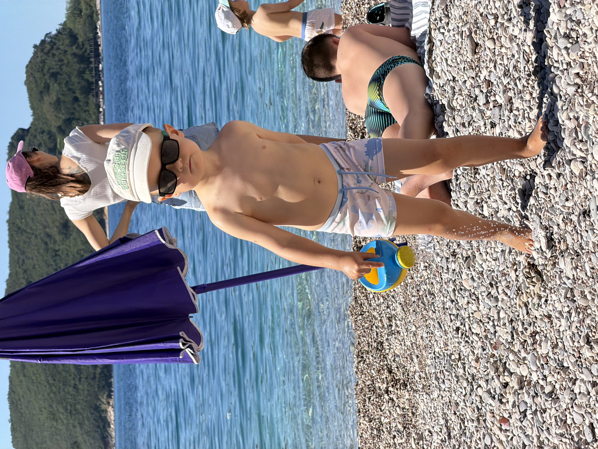 The image captures a sunny scene on a pebble beach, most likely in Boreti, Montenegro, given the clear turquoise water and mountainous backdrop. The scene is dominated by a young boy wearing a white cap and sunglasses, holding a blue and yellow water can. He is standing on the beach near the water's edge. Adjacent to him, to the left, is a purple beach umbrella. In the background, there are two more children and a woman in a pink hat, seemingly part of the same group. One child is lying on the beach, and the other is standing near the water. The beach is covered in light-colored pebbles, and the water is clear with minor ripples. The background features lush green mountains under a clear sky.