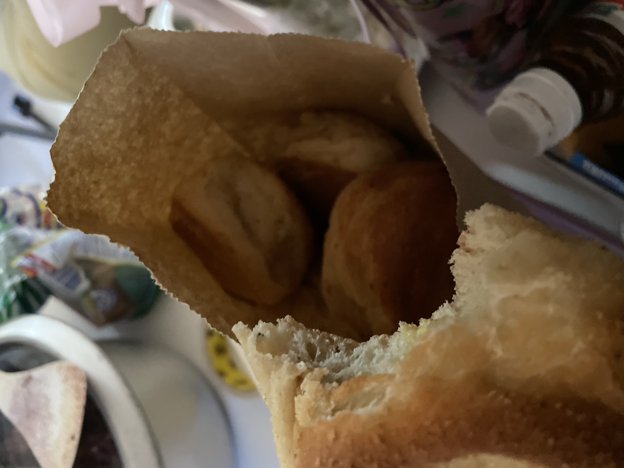 A close-up shot captures a brown paper bag filled with small, golden-brown rolls. A portion of a larger, torn bread with a soft, white interior is visible in the foreground, partially obscuring the bag. The background is blurred, showing hints of a white bowl with a reddish substance, a yellow object, and various packaging materials. The scene appears to be indoors, possibly in a kitchen or dining area. No specific location in Davao City, Philippines can be determined from the image.