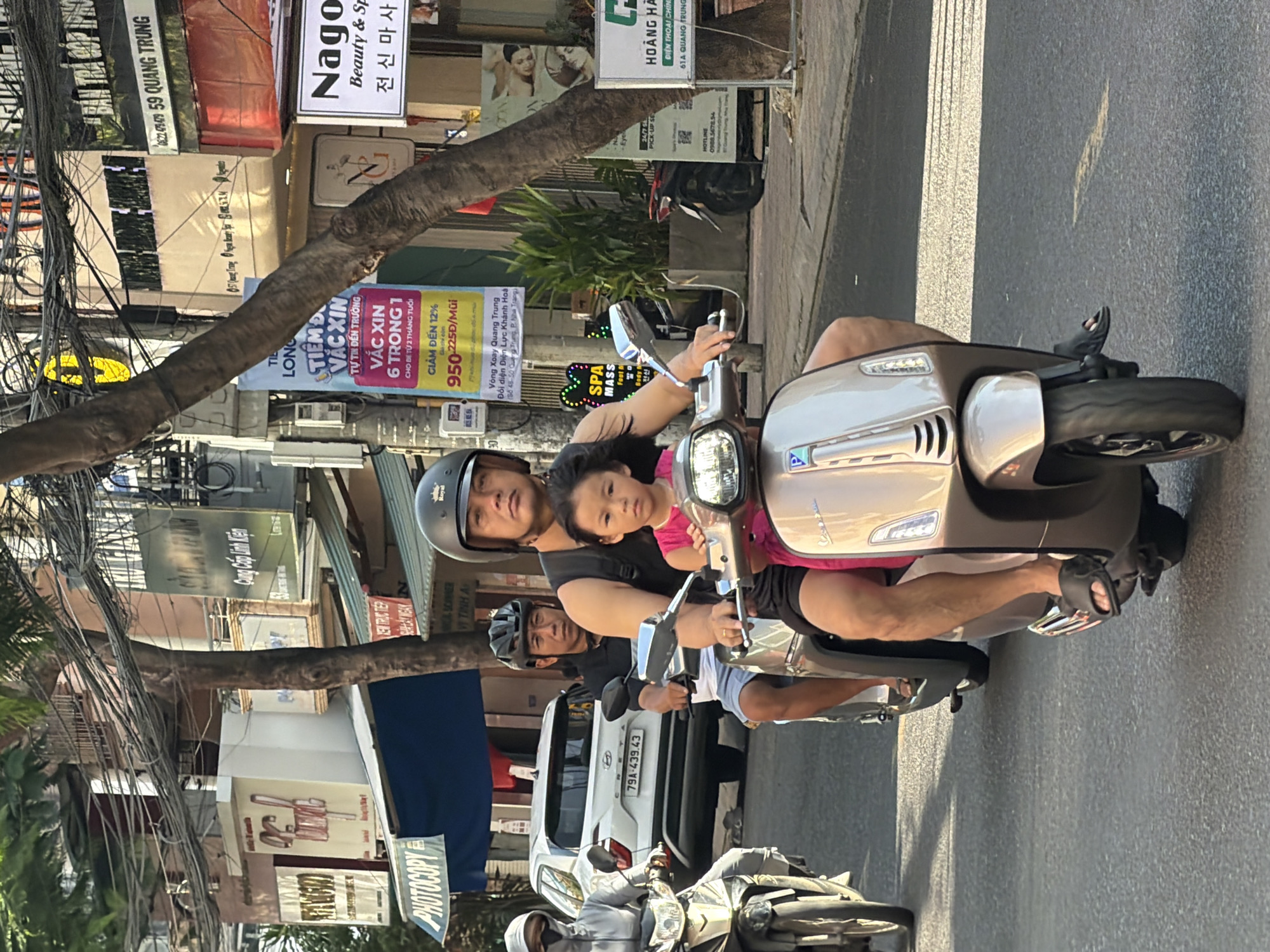 A medium shot shows three people on two motorbikes on a street in Nha Trang, Vietnam. On the left, a person wearing a grey hooded jacket and a mask rides a white motorbike. On the right, a man wearing a black helmet and a black sleeveless shirt rides a tan Vespa scooter, with a young girl in a pink dress sitting in front of him. Behind the man on the Vespa, another man in a black shirt and helmet rides a black motorbike. Several buildings with signs in Vietnamese are visible along the street. The sky is clear and sunny.