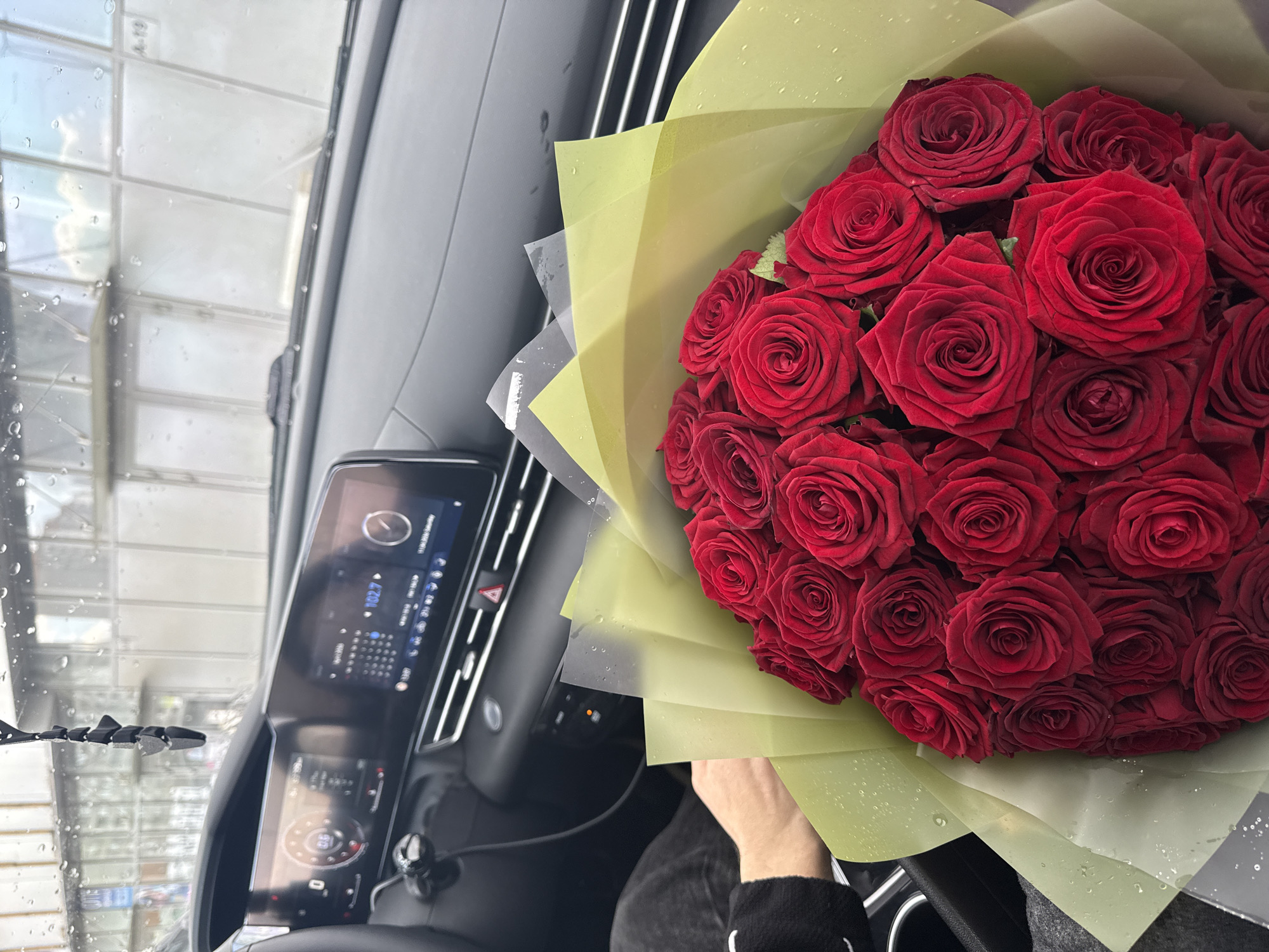 The photograph shows a close-up view from inside a car. On the right, a large bouquet of red roses wrapped in light green paper is held by a hand wearing a black sleeve. The car's dashboard is visible on the left, featuring a digital display with readable radio station information. Through the car's windshield, there is a view of what appears to be an automated car wash. The location is unknown.