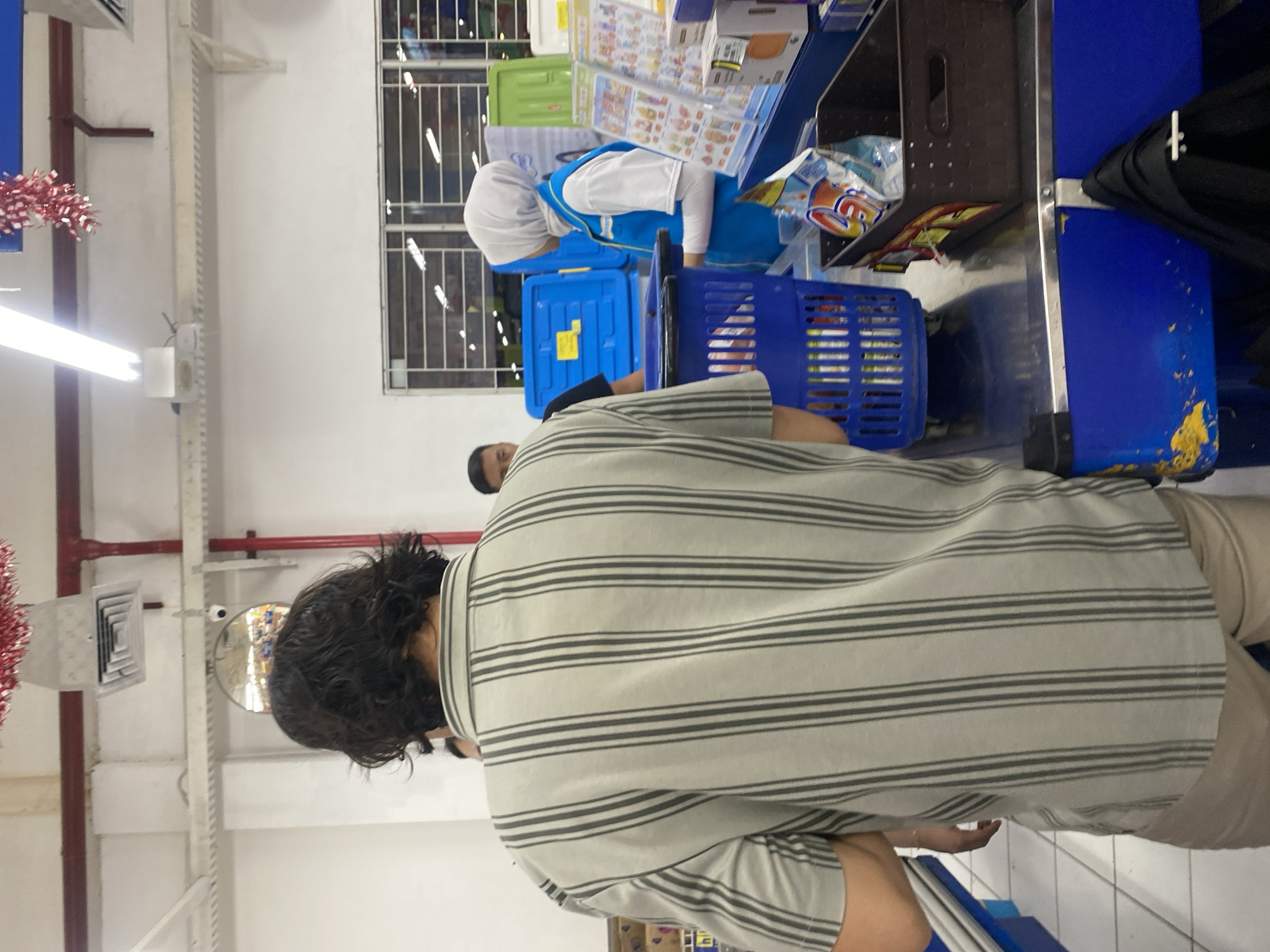 The image depicts a checkout counter in a supermarket, likely located in Banjarbaru, Indonesia. A person, viewed from behind, wearing a grey and black striped shirt and tan pants, stands at the counter. Next to them, a blue shopping basket is filled with items. A store employee wearing a blue vest and white hijab is attending to the checkout. Other individuals are visible in the background, including a man in a black t-shirt and another man partially obscured. The environment appears to be a typical retail setting with bright overhead lighting and shelving units visible in the distance. The checkout counter is blue and silver, with a dark brown bin containing grocery items.