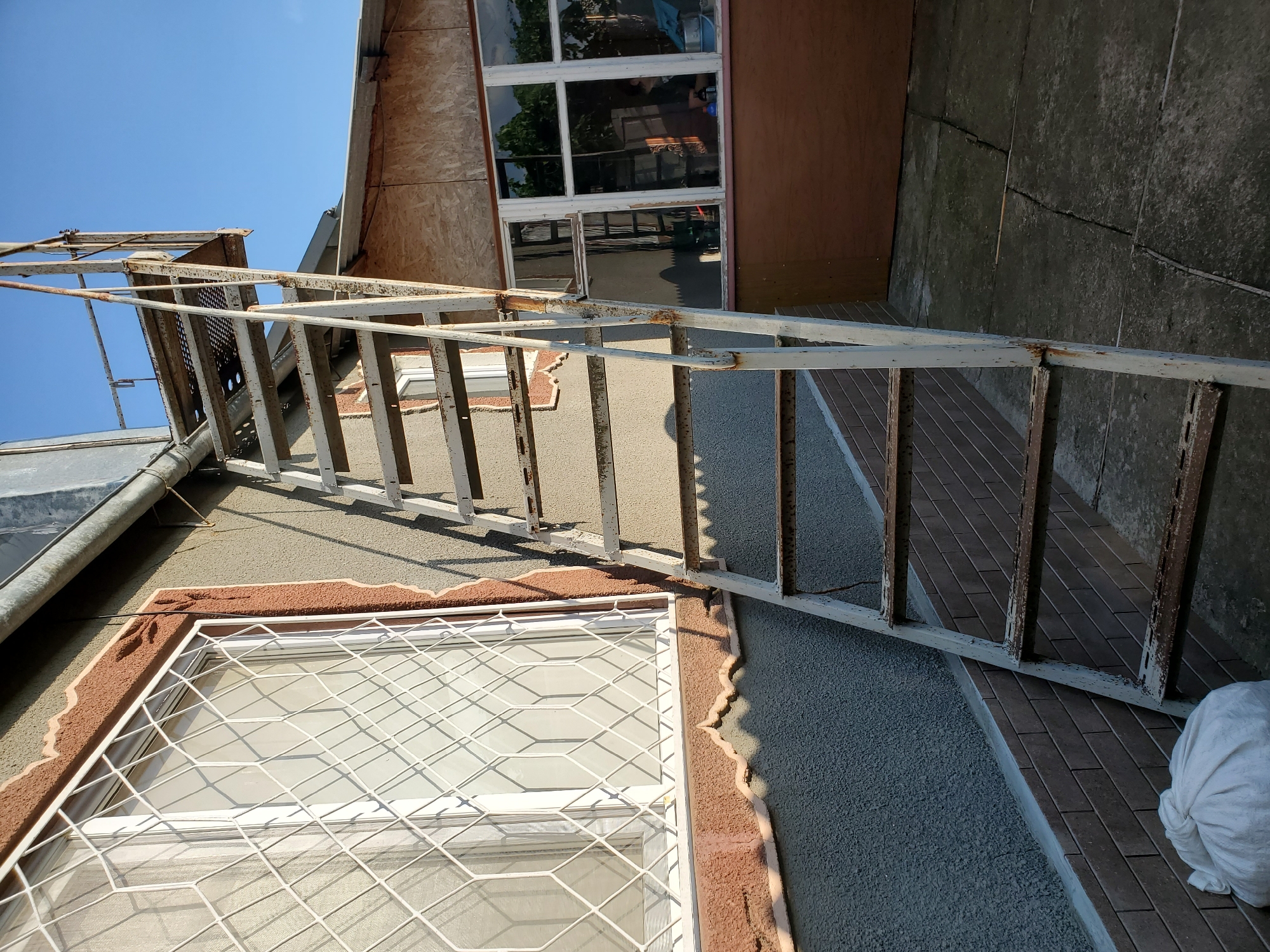 An outdoor image shows a rusty, white metal staircase leading up the side of a building with a gray stucco exterior and a brown textured trim around a window. The window has a white diamond-patterned grate. Adjacent to this is a window with a light brown frame, reflecting trees and a blue sky. A section of the building is covered with light brown plywood. The ground is paved with cracked concrete slabs. A white sack is visible at the base of the wall, near a section of dark brown brickwork. The sky is clear and blue. The scene appears to be in Konstantinovka, Moldova.