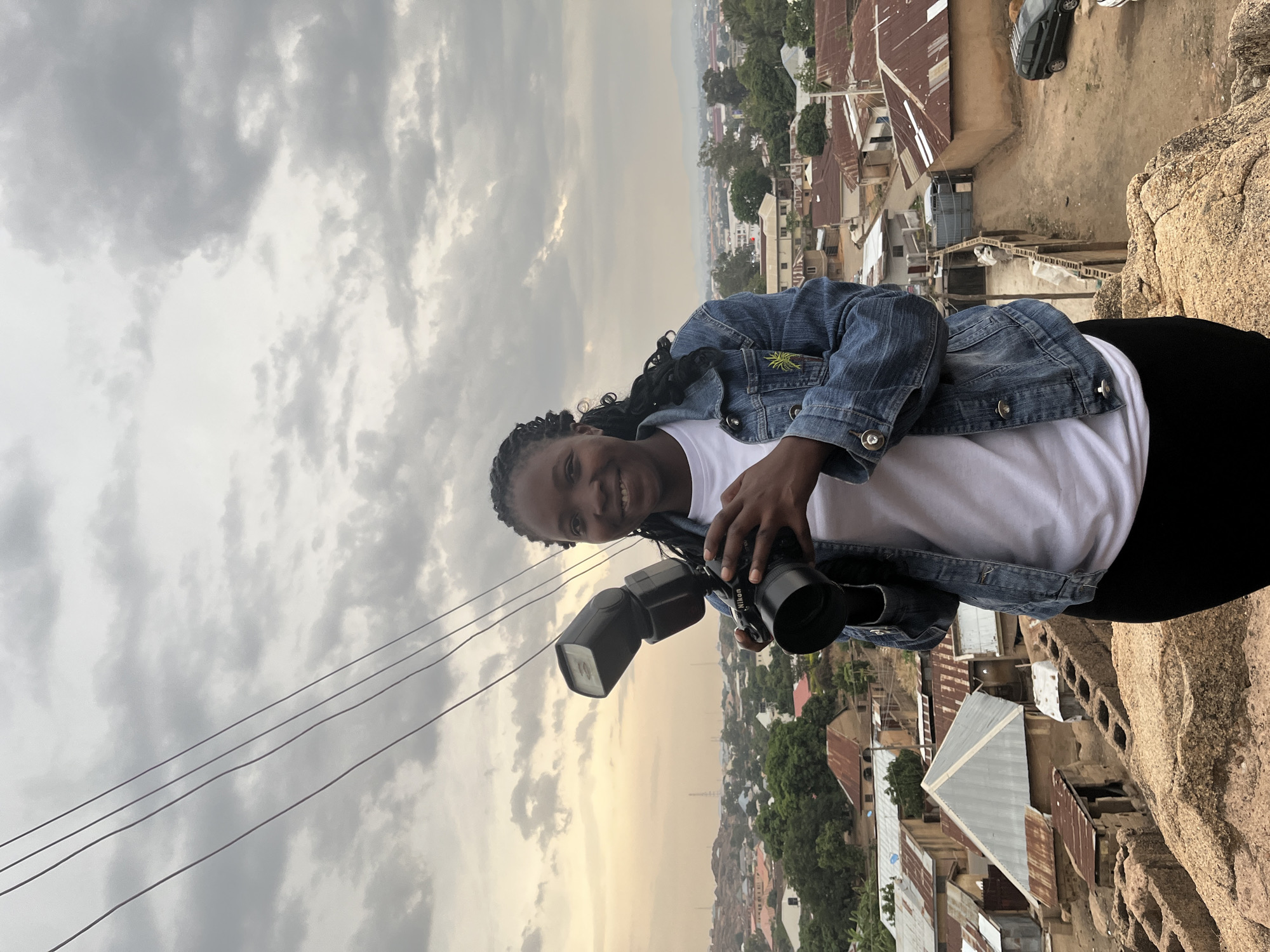 A smiling woman in a denim jacket and white t-shirt stands on a rocky outcrop, holding a Nikon camera with a mounted flash. She is outdoors, overlooking the city of Jos, Nigeria, identifiable by the mix of buildings with corrugated metal roofs and lush greenery. The sky is overcast with grey and white clouds. Power lines are visible in the upper left corner.