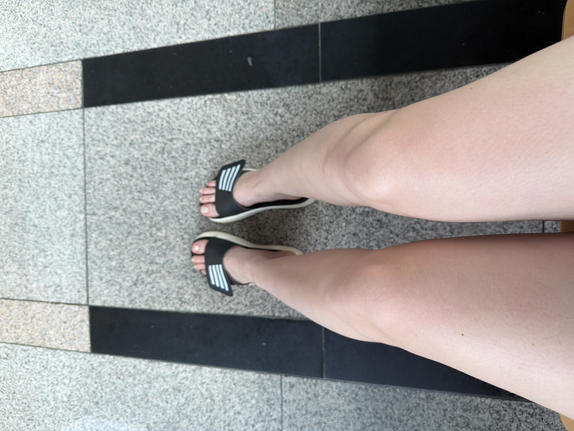 A person's legs and feet are shown from a top-down perspective, sitting on a bench. They are wearing black slide sandals with white stripes. The background is a textured stone-like floor with black and grey rectangular sections. The scene appears to be outdoors.