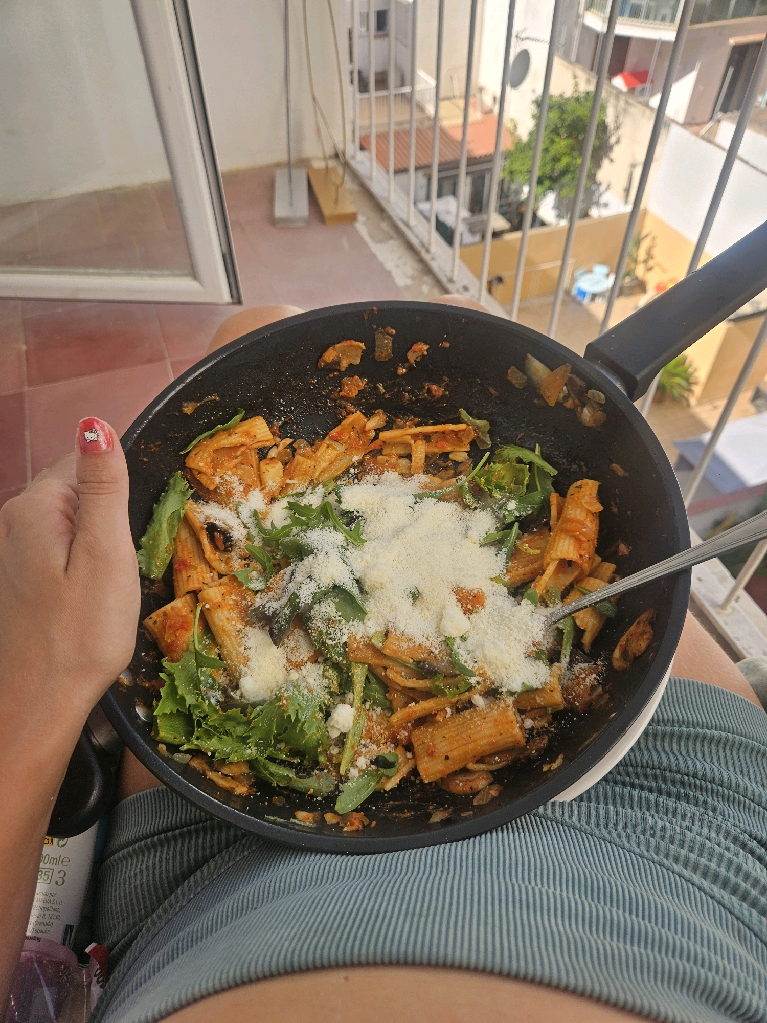 A person is holding a black frying pan filled with rigatoni pasta in a red sauce, topped with fresh green herbs and grated cheese. The person's thumb, adorned with a Hello Kitty nail sticker, is visible on the left side of the pan. A fork rests in the pasta. The scene is set on a balcony overlooking a cityscape, likely in Palma de Mallorca, Spain, suggested by the architecture with white facades and terracotta roofs, and the outdoor furniture visible in the background. The floor is tiled in a reddish-brown hue. The person is wearing light blue striped shorts.