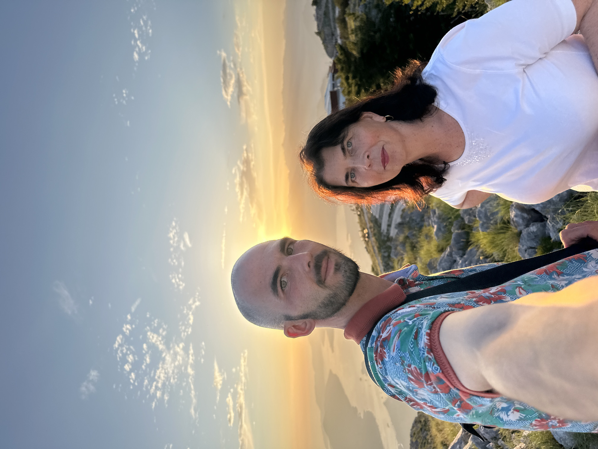 This is a selfie taken by a man in the foreground, with a woman beside him. They are standing on a rocky, grassy slope with a panoramic view of a bay and mountains in the background. The sun is setting, casting a warm golden light across the scene and illuminating the sky with soft clouds. The man is wearing a colorful, patterned shirt, and the woman is wearing a white t-shirt. The location is identified as Škaljari, Montenegro.