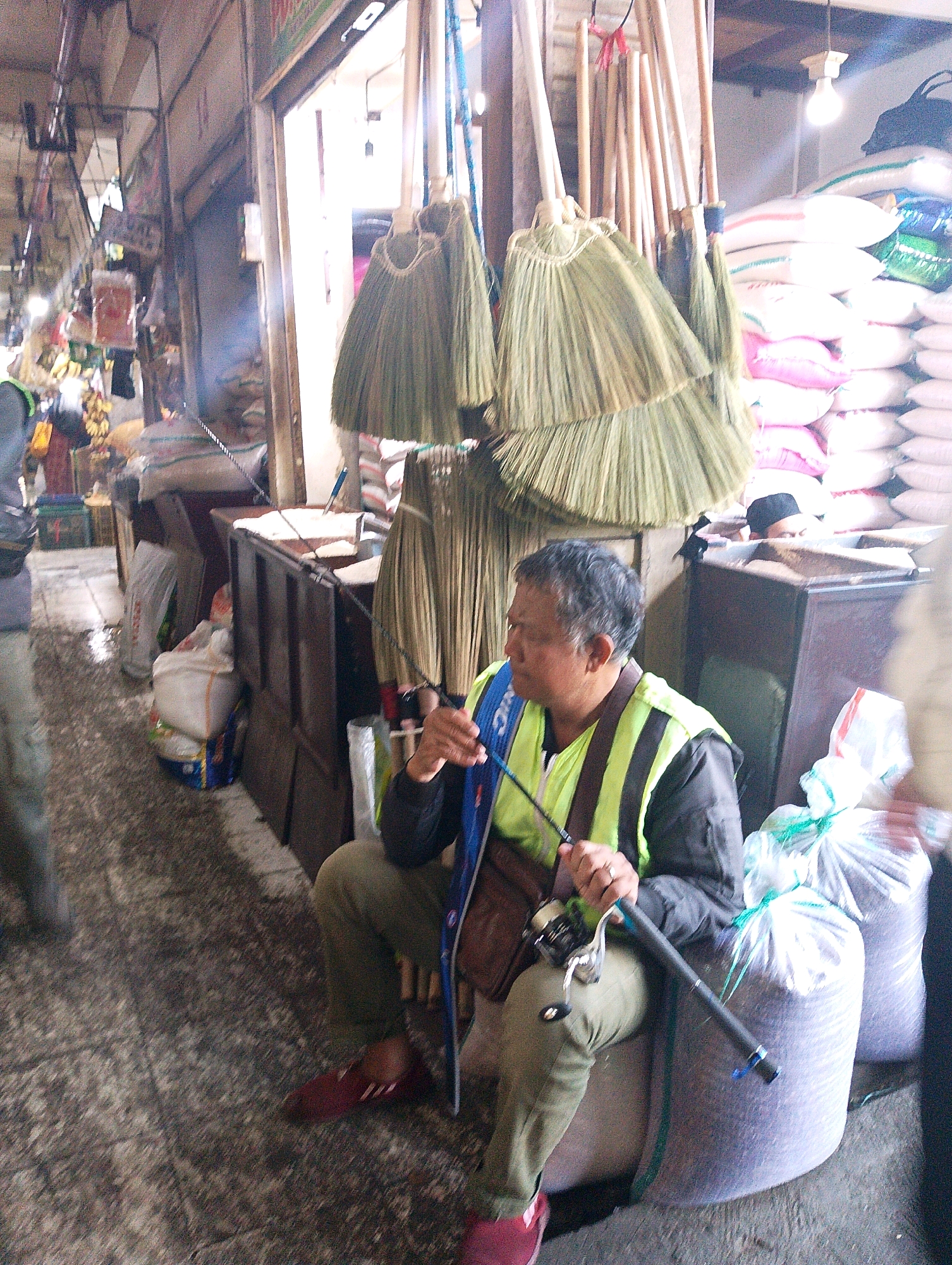 In Lembang, Indonesia, within an indoor market, a man is seated on a large sack of grain. He is wearing a bright green vest with black stripes over a dark jacket and olive-colored pants, along with maroon shoes. He holds a fishing rod with a reel, its tip extending towards the left side of the frame. The background reveals stacks of similar sacks, some pink and white, and an assortment of brooms hanging from above, suggesting a marketplace setting. The floor appears wet and gritty, indicative of a busy, possibly outdoor or semi-outdoor, market environment.
